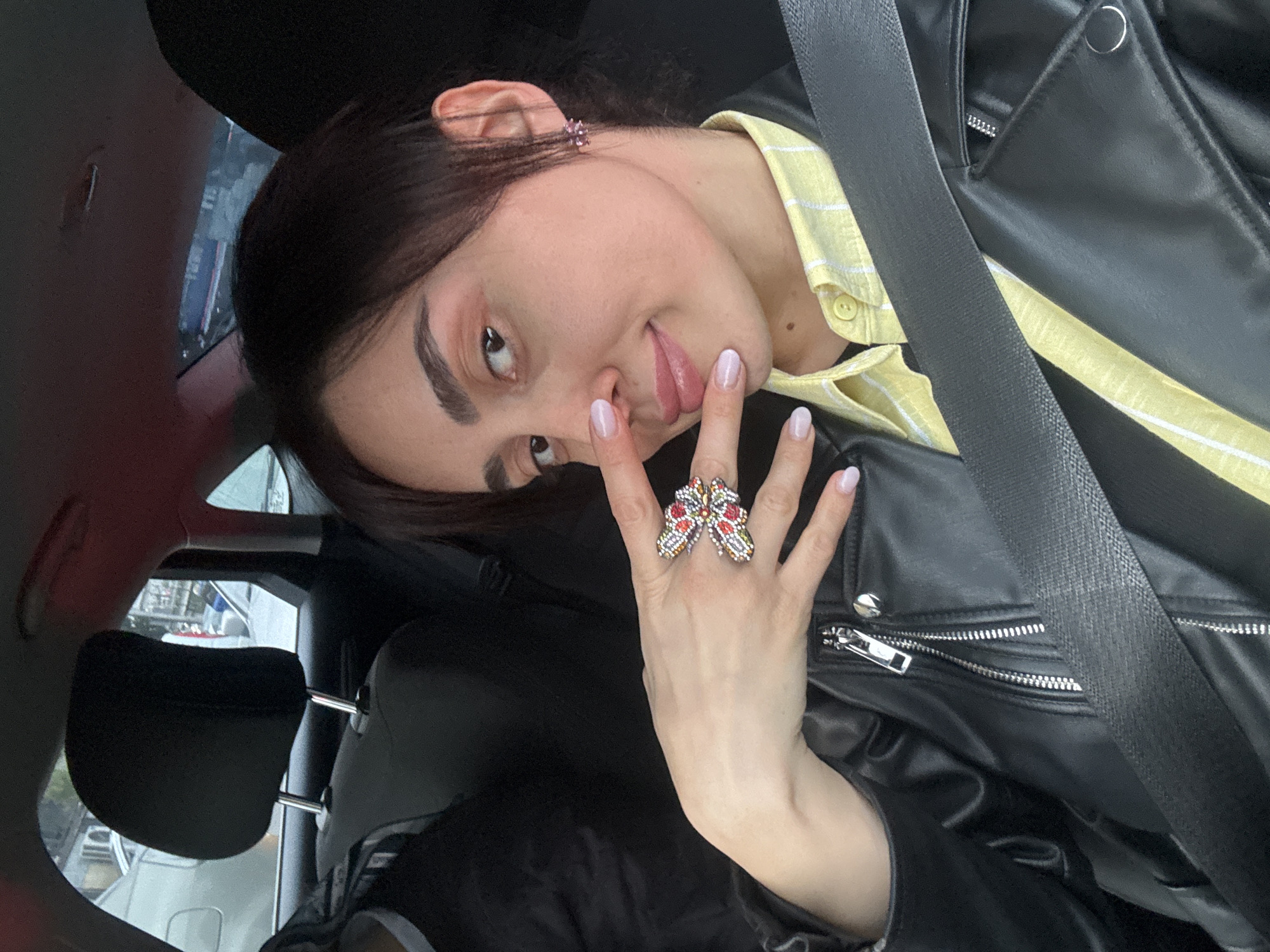A young individual is captured in a close-up portrait, oriented sideways in the frame. The person, likely female, has dark hair and pronounced dark eyebrows. They are looking directly at the camera with a playful expression, sticking their tongue out slightly while their right index finger touches their nose.
They are wearing a black faux leather jacket with silver zipper details and a snap button, over a light yellow shirt with thin white stripes. A black seatbelt crosses their chest, indicating they are in a vehicle. On the ring finger of their right hand, they wear a large, colorful butterfly-shaped ring adorned with red, yellow, and orange beads or stones. A small, purple, floral-shaped earring is visible on their right ear. Their nails are manicured and painted with a light, possibly clear or pale pink, polish.
The setting appears to be the interior of a car. A black headrest is visible behind them, and through the car window, blurred elements of an urban environment, possibly other vehicles and buildings, can be discerned, consistent with a daytime scene. The overall mood is casual and lighthearted. There is no visible text in the image. The location context provided is Tbilisi, Georgia.