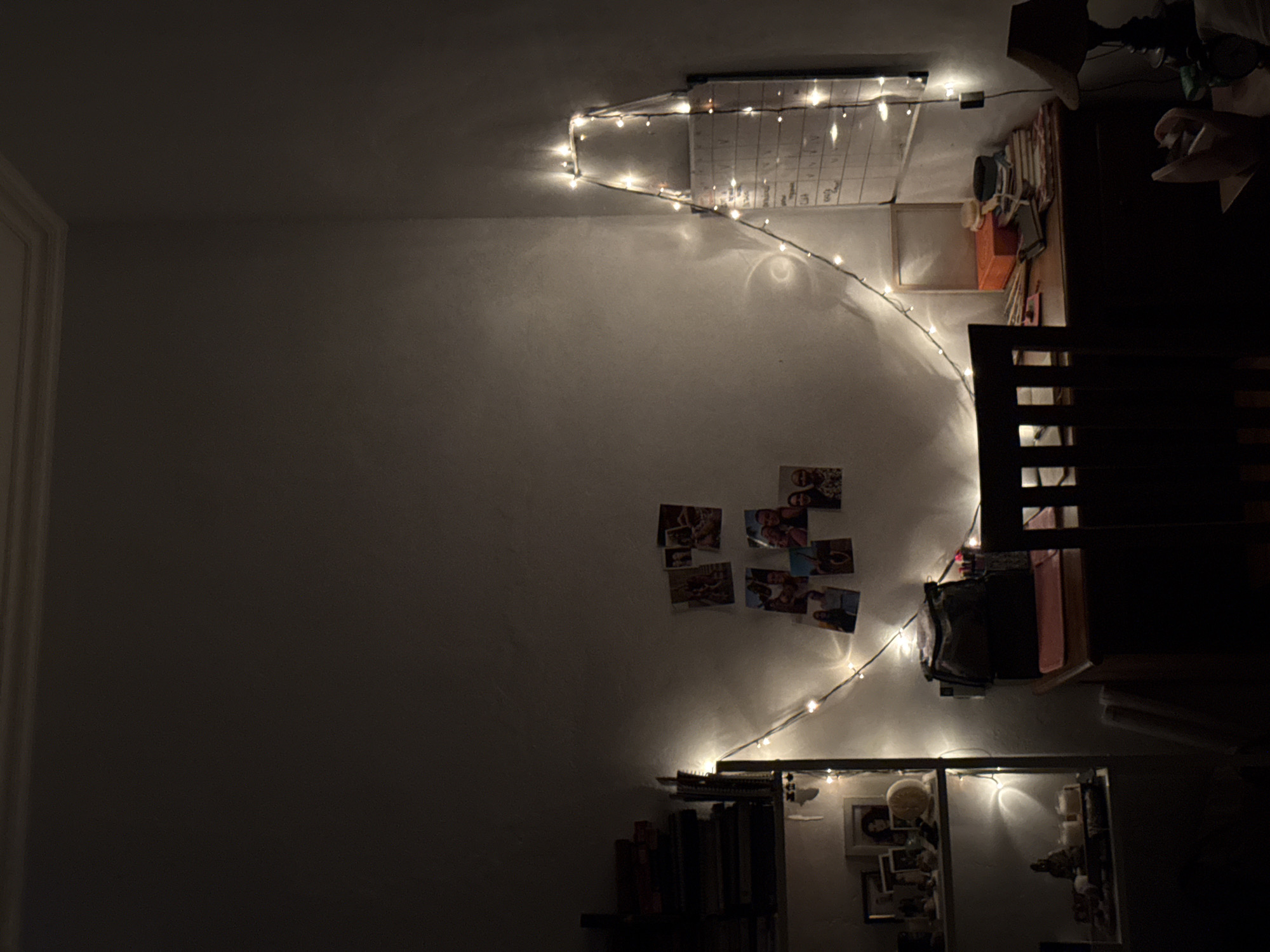 The image depicts a room interior illuminated by string lights. The left side of the room is dark, leading to a wall with a string of lights arched across it. Several photographs are affixed to the wall. A calendar hangs near the ceiling, also adorned with lights. To the right, a partially obscured desk and chair are visible. Below the photos, a bookshelf is present, with books and framed pictures. The photograph does not show any evidence to indicate it was taken in Tampaksiring, Indonesia.