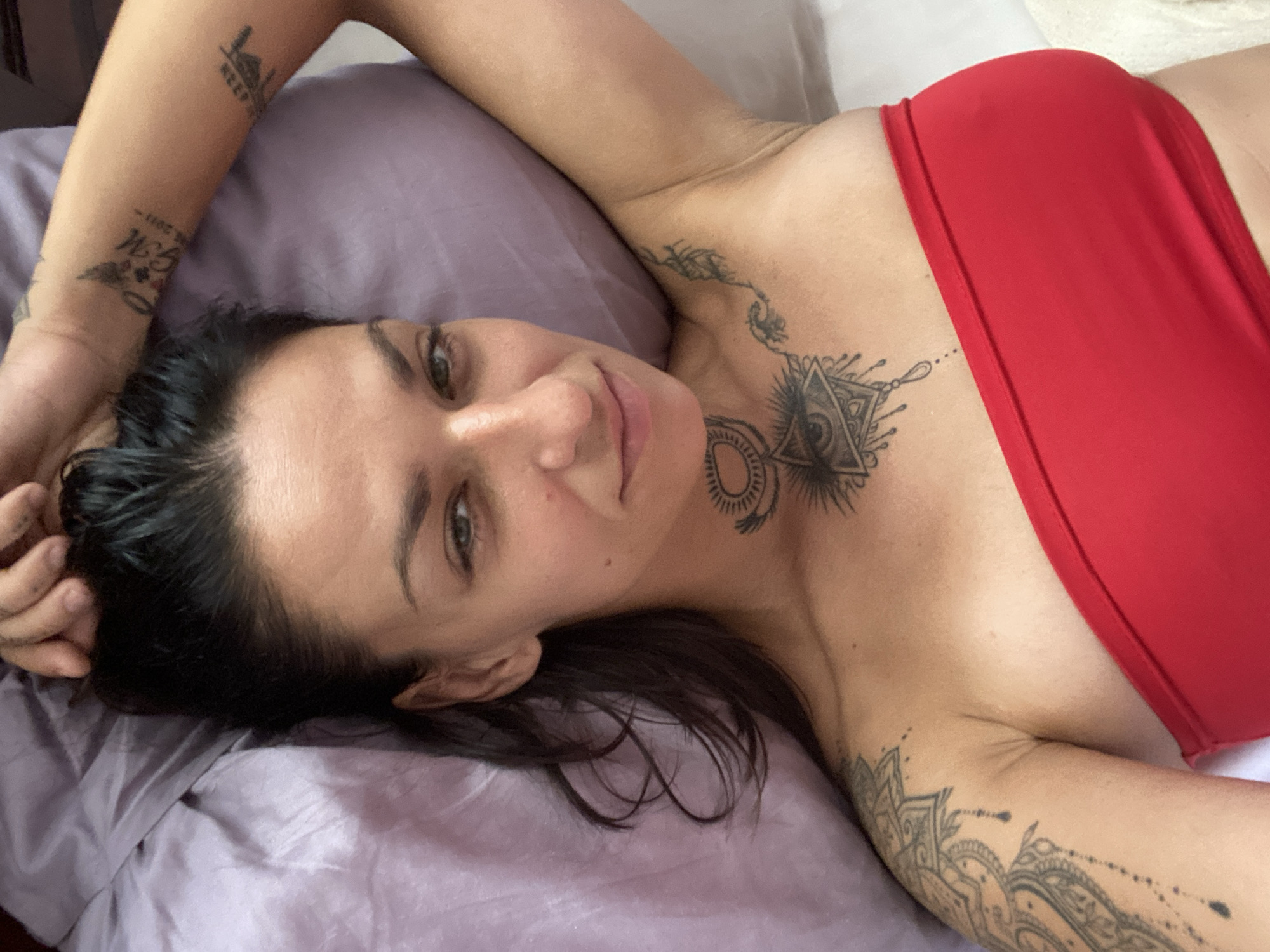 A woman with dark hair and green eyes lies on a lilac-colored pillow in a close-up, overhead shot. She is wearing a red strapless top and has several tattoos visible on her arms, chest, and neck. The tattoo on her chest resembles an eye within a triangle with sun rays. She is looking directly at the camera with a slight smile. The background consists of the pillow and what appears to be a white surface. There is no visible indication of the city of Mũi Né, Vietnam, in the image.