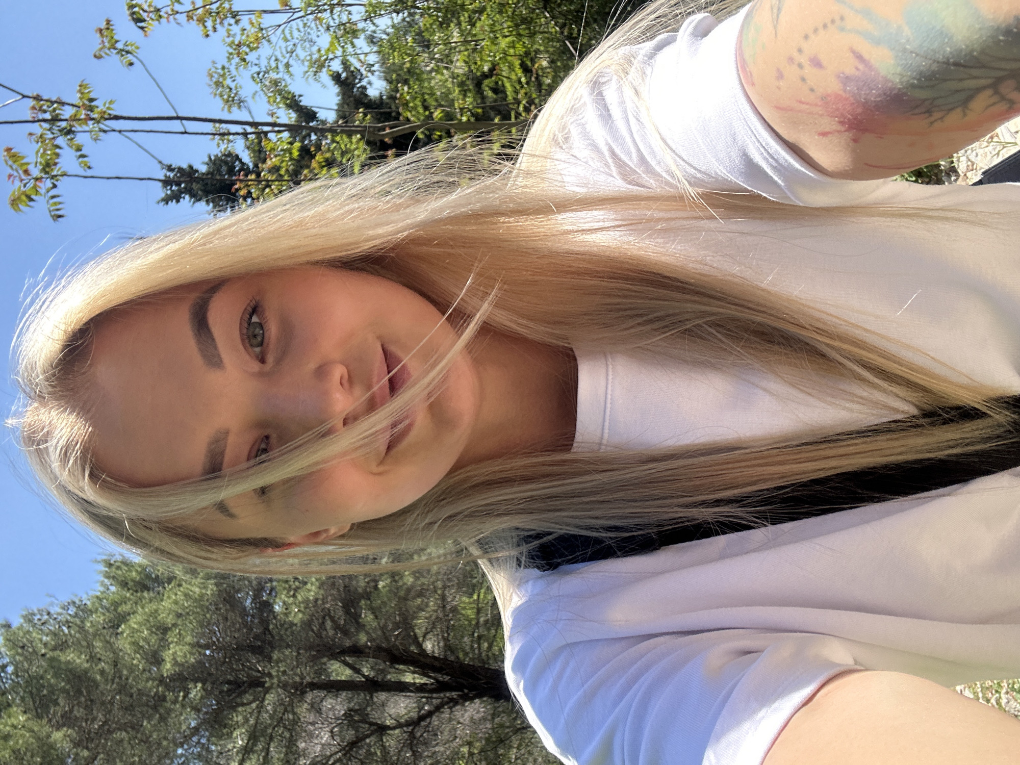 A fair-skinned female with long, blonde hair is shown in a selfie outdoors. The background includes trees and a clear blue sky. The female is wearing a white shirt, and part of a tattoo is visible on her upper left arm. The location is possibly in or near the city of Budva, Montenegro.