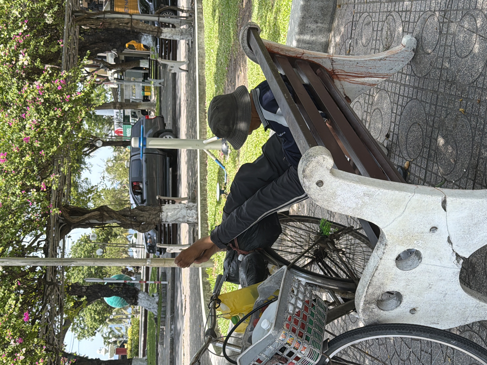 In Nha Trang Ward, Vietnam, a person is seen resting on a park bench, with their legs extended and propped up on a bicycle. The bicycle has a basket filled with various items, including plastic bags and bottles. The person is wearing a dark bucket hat and dark clothing. The bench is made of wood and concrete, with some reddish staining on the leg. The scene appears to be outdoors in a public area, with trees and flowering vines overhead, and cars visible in the background. The ground is paved with tiles.
