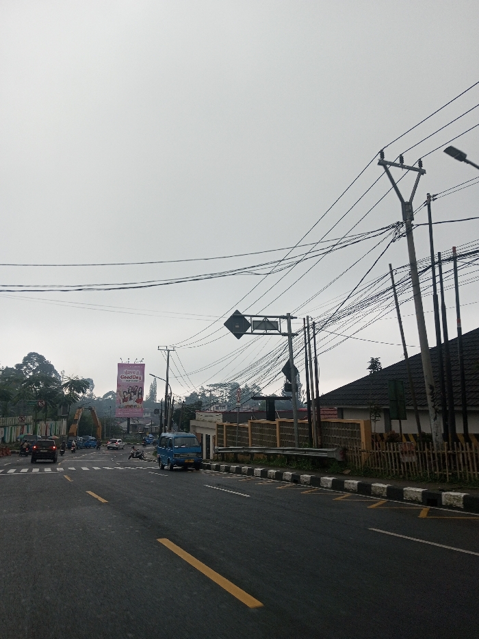 The image captures a detailed street scene under an overcast sky, likely in an urban or semi-urban area of Indonesia, suggested by the visible text and vehicle type. The time of day appears to be daytime, but with diffused light due to the heavy cloud cover.
In the foreground, a dark asphalt road dominates the lower portion of the image. It features clear yellow double solid lines, indicating a no-passing zone, and a single dashed yellow line, likely for a turn lane or parking. On the right side of the road, a black and white striped curb with yellow markings suggests a restricted parking or stopping area. A small, light blue van, possibly an angkot (public minivan), is stopped or parked adjacent to this curb.
Further down the road, in the midground, several vehicles are visible. A red car is prominent on the left side of the road, with other cars and motorbikes visible behind it, suggesting active traffic flow or an intersection. To the far left, a partially visible green and white sign reads "PUNCAK" followed by obscured text like "MENUJU..." and "TERBUKA UNTUK". This strongly suggests the location is near the Puncak area in West Java, Indonesia. Behind this sign and the vehicles, dense green foliage indicates a treed or hilly landscape.
A large, brightly lit billboard stands tall on the left side of the road, featuring an advertisement for "GoodDay" coffee, with the words "Kopi Gaul" also visible below it. The billboard depicts several individuals, likely enjoying the beverage. Below the billboard and partially obscured by trees, some construction equipment, possibly an excavator or crane, can be seen.
The midground and right side of the image are characterized by a dense network of overhead utility wires and numerous utility poles. One prominent pole near the center of the image supports two diamond-shaped road signs, one directly above the other, though their specific meaning is not clear from the angle. Another large utility pole with multiple cross arms and wires stands on the far right.
Behind the utility poles and wires on the right, there is a building with a dark, possibly tiled, roof. A fence made of light-colored blocks and wood runs along the property line, separating the building from the road. Adjacent to the blue van, a small, light-colored building or guard post is visible. The background shows more distant hills or elevated terrain covered in trees, blending into the consistently overcast sky.
No specific activity beyond general traffic movement is evident, and no distinct emotions are discernible from the scene. The image presents a typical, bustling road environment in a Southeast Asian setting.
