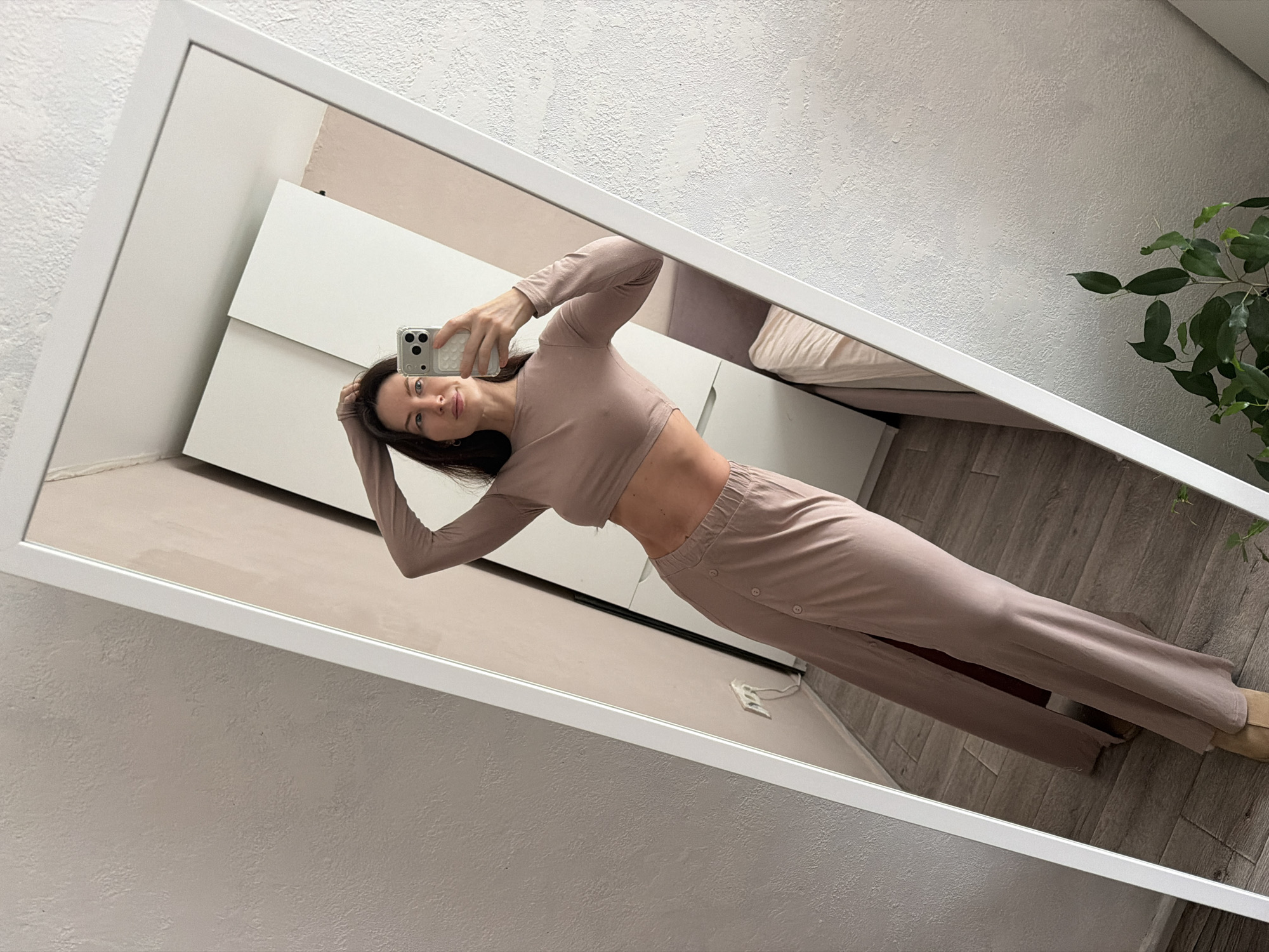 A young woman is taking a mirror selfie in what appears to be an apartment in Perm, Russia. She is wearing a light brown two-piece outfit consisting of a long-sleeved crop top and pants with buttons on the side. She is holding her phone in her right hand and has her left hand on her head. The mirror reflects a room with white furniture, a bed, and a plant. The walls are a light color.
