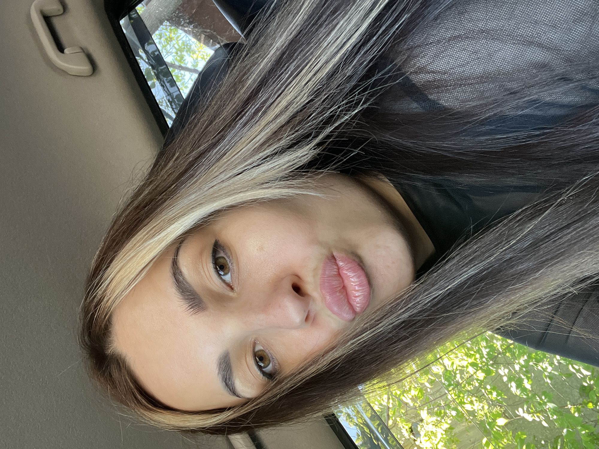 A selfie photograph shows a young woman with brown eyes and highlighted brown and blonde hair. She is in a car, likely a passenger, judging by the view of trees and sky through the window on the right and the car interior on the left. She is looking directly at the camera with slightly pursed lips. The city is Unknown, Unknown.