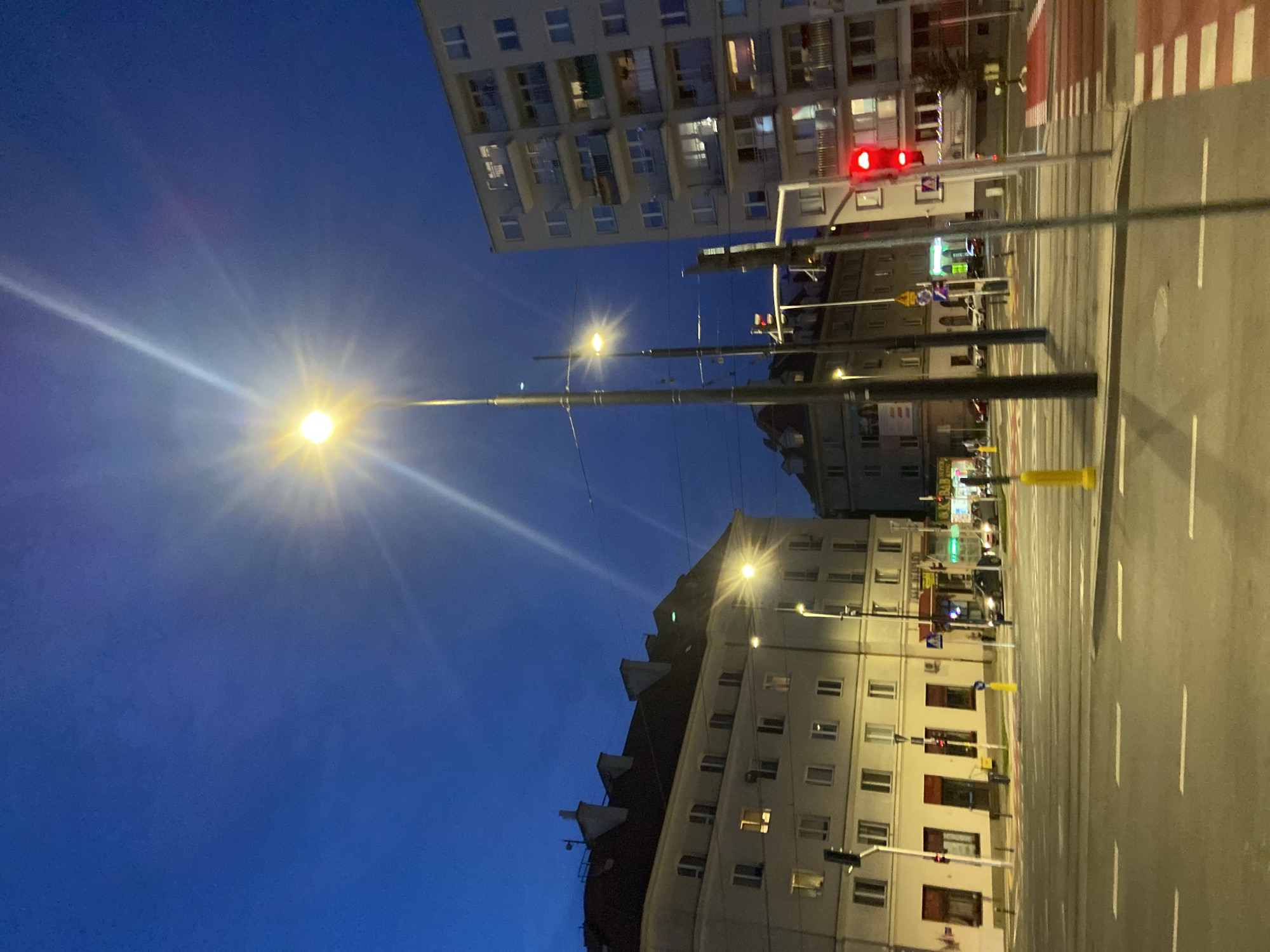 The image captures a street scene in Warsaw, Poland, at dusk. A bright street lamp in the foreground casts a strong glare, creating a sunburst effect against the deep blue twilight sky. To the left, a tall, modern apartment building with balconies and numerous windows is visible. Another older, multi-story building with a dark red tiled roof and dormer windows stands to the far left. A traffic light displaying a red signal is prominent on the right side of the frame, illuminating a red-and-white striped pedestrian crossing. Several cars are present on the wet asphalt road, suggesting recent rain or ongoing street cleaning. The urban environment appears to be a busy intersection, with streetlights and building lights illuminating the area. The overall atmosphere is that of a city transitioning from day to night.
