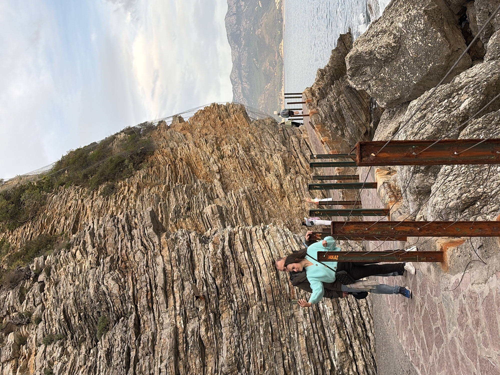 The image captures a scene at a coastal path, likely the Sveti Stefan section, near Budva, Montenegro. A couple is walking along a pathway constructed with rusted metal railings and stone-paved ground. The woman, wearing a turquoise sweater and black pants, is holding a smartphone, suggesting she might be taking a picture. The man is walking next to her.
The path runs alongside a steep, rocky cliff face with distinct geological layering. A safety net is secured along the upper edge of the cliff to prevent rockfall. The sea is visible in the background, along with distant mountains. Other people can be seen further along the path. The sky is overcast with light, diffused sunlight.
