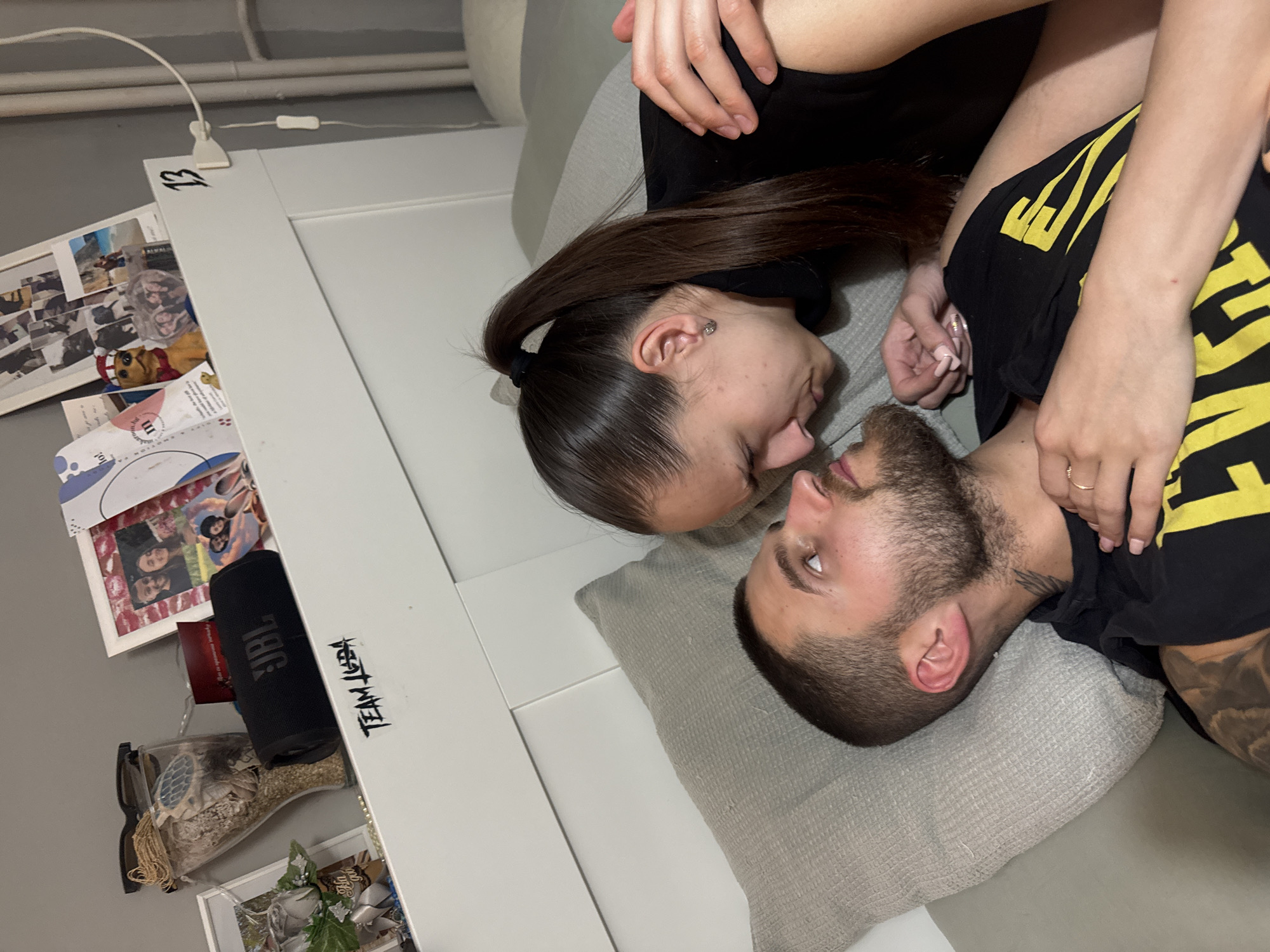 This image captures an intimate indoor moment between a man and a woman, likely on a bed or sofa in Sofia, Bulgaria. The woman, with dark hair tied back in a ponytail and wearing a black top, is positioned above the man, looking affectionately into his eyes. Her left arm is gently draped over his shoulder, and her fingers, adorned with light pink nail polish, rest near his neck. She wears a small earring in her left ear.
The man, with short dark hair and a neatly trimmed beard, is lying on his back, gazing up at the woman. He is wearing a black t-shirt with yellow lettering or design visible on the front, and a tattoo covers a portion of his left arm. A small ring is visible on his right hand. Both individuals are resting their heads on grey textured pillows.
To the left of the couple, a white headboard or frame serves as a shelf, displaying various personal effects. These include multiple framed photographs, some appearing to be collages, and a small yellow dog toy wearing a red and white Santa hat. A black JBL portable speaker stands upright next to the photos. Further down, a clear glass jar filled with sand and seashells, and a small artificial plant arrangement, are visible. The wall in the background is a plain grey.
Visible text includes the number "13" marked on the white frame near a white clip holding a wire. "JBL" is clearly printed on the speaker, and "TEAM LEBA" is written vertically on a sticker or label attached to the white frame. The overall atmosphere conveys a sense of closeness and comfort between the two individuals.