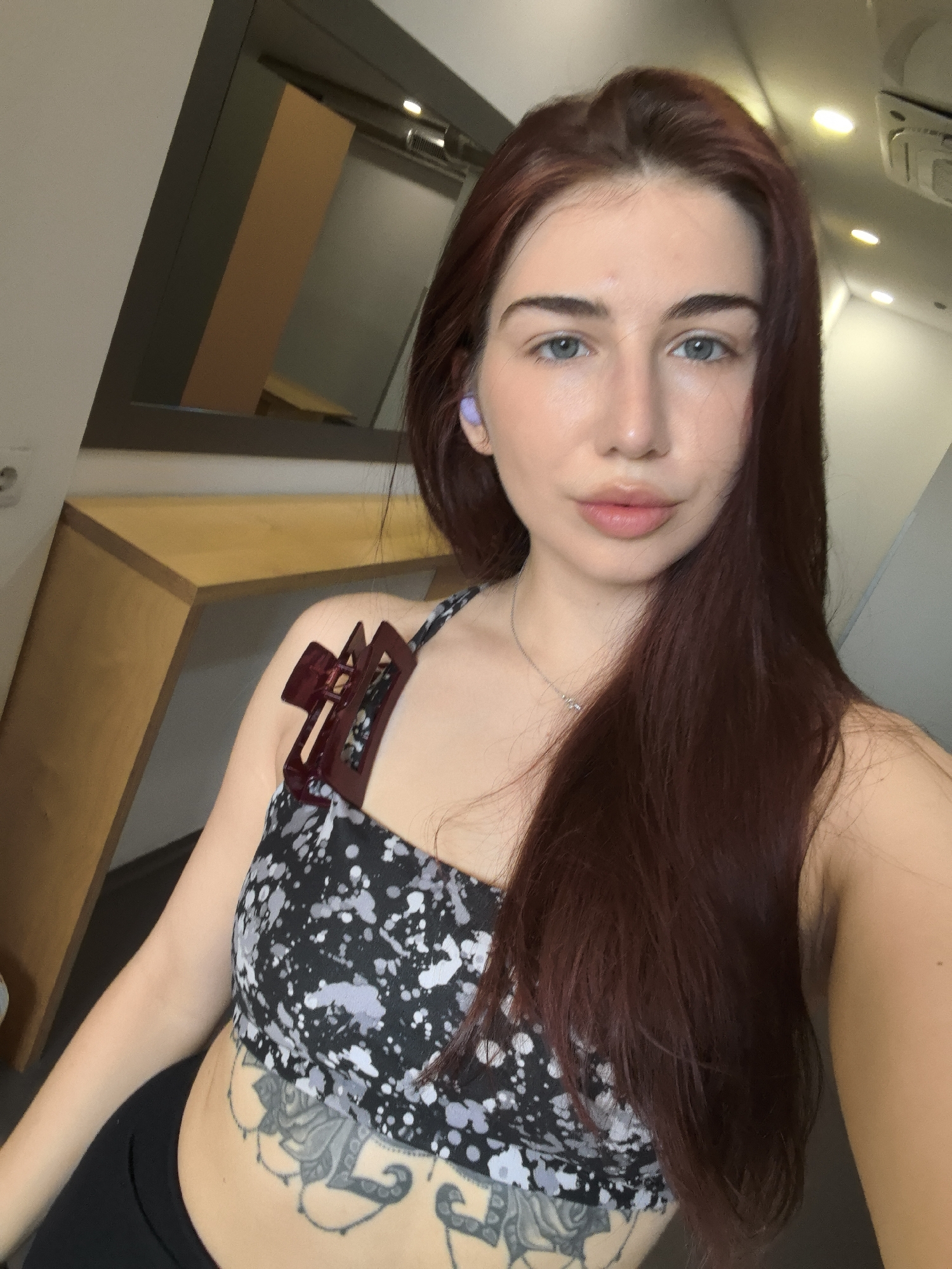 A woman with long, auburn hair is taking a selfie indoors. She is wearing a black and white patterned tank top, black pants, and a necklace. She has a tattoo visible on her abdomen. A maroon hair clip is attached to the strap of her tank top. She has a lilac-colored earbud in her left ear. The background includes a partially visible table or counter and a mirror reflecting the room's interior. The environment appears modern and well-lit with overhead lighting. There are no indicators that suggest the location is in Stara Zagora, Bulgaria.