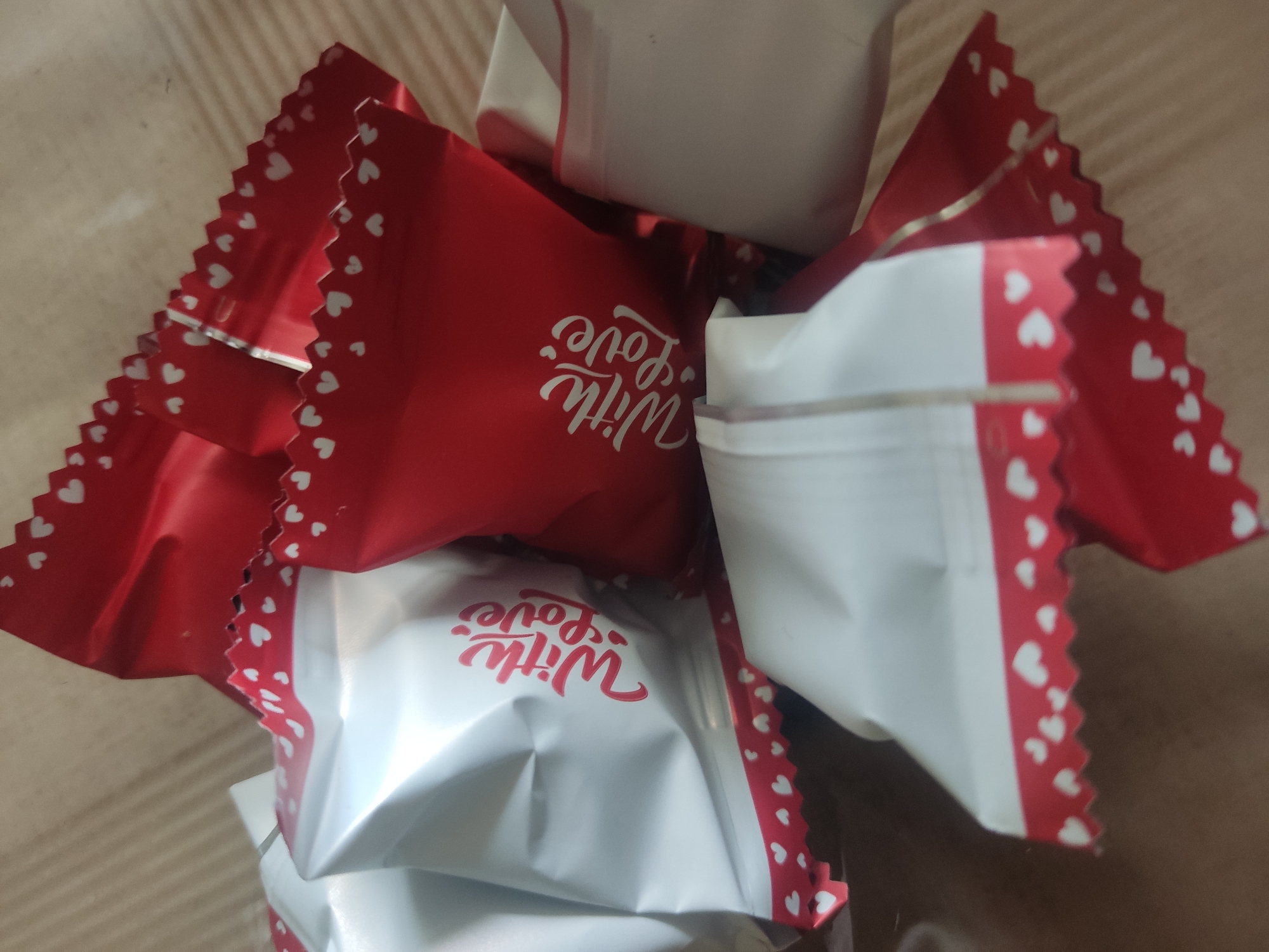 A pile of chocolates individually wrapped in red and white foil is scattered on a wooden surface. The foil wrappers are decorated with small white hearts and a scalloped edge. Some of the wrappers have the words "With Love" printed in a script font in red or white. The lighting is overhead, creating soft shadows. The image is an extreme close-up, with no background features visible beyond the chocolates and the surface they are on. The location is unknown.
