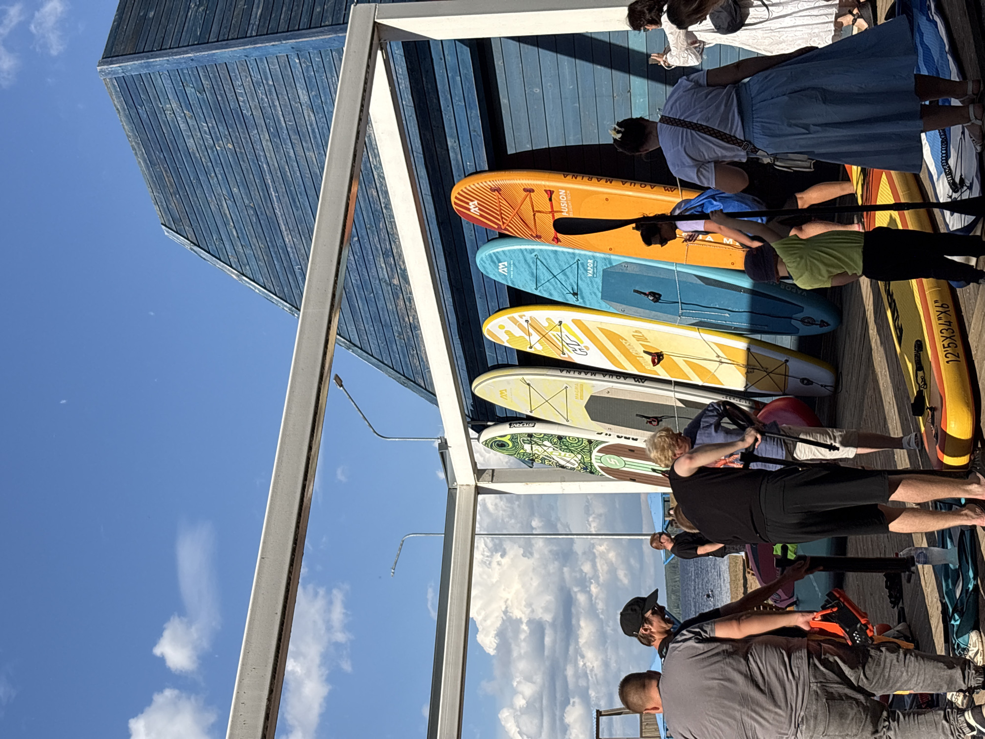 In Chaikovsky, Russia, on a sunny day, a group of people stand on a wooden pier next to a row of colorful paddleboards leaning against a blue wooden building. The paddleboards are of various colors including green, yellow, blue, and orange, with some displaying brand names like "Aqua Marina." Several individuals are visible in the foreground and midground, some wearing casual clothing such as t-shirts, shorts, and jeans. One person in the foreground is holding an orange paddleboard accessory. In the background, a body of water can be seen, with a single person on a paddleboard in the distance. The sky is blue with scattered white clouds, and a portion of a white canopy structure is present in the foreground.