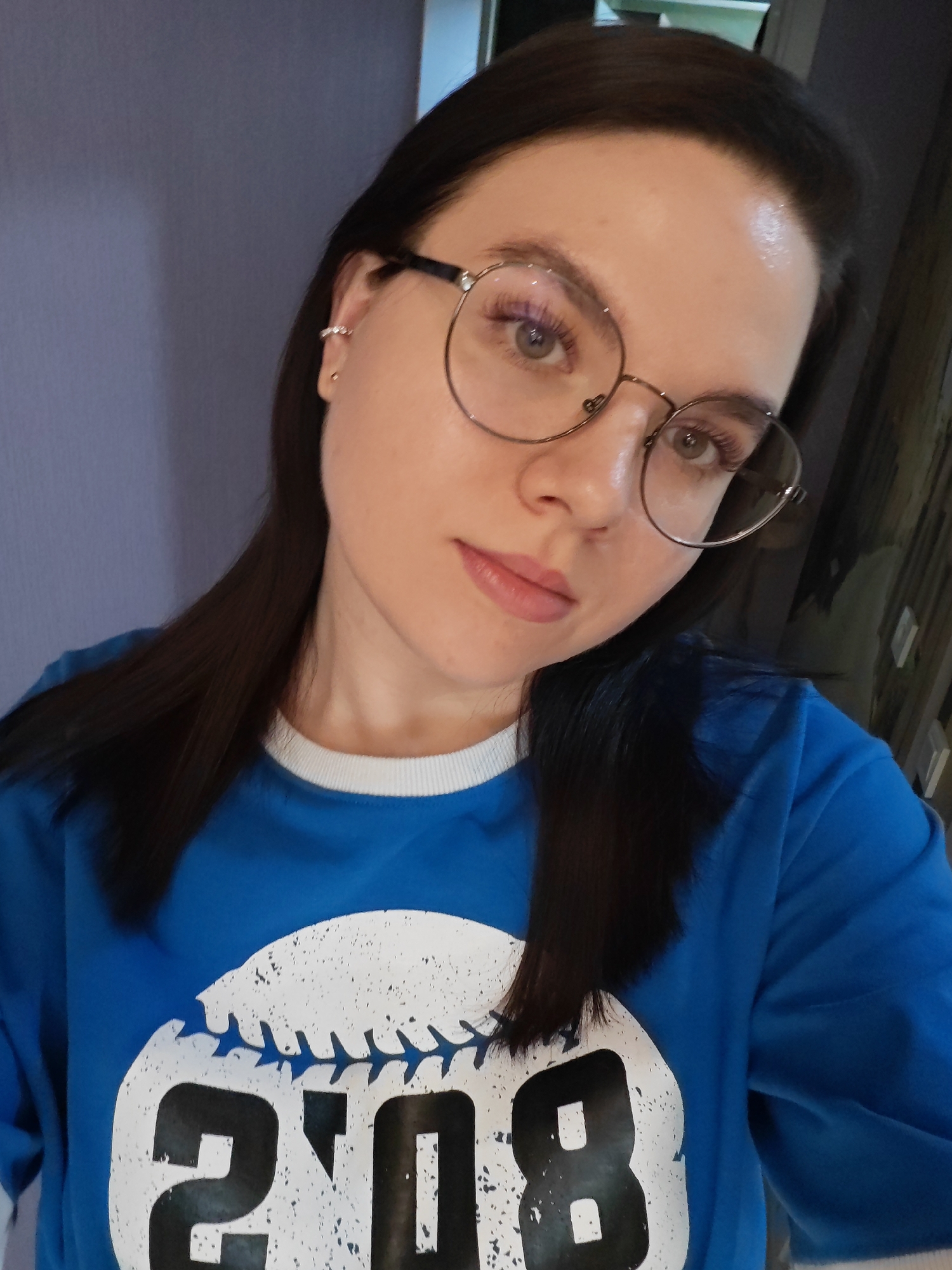 The image shows a close-up of a young woman with dark hair and glasses, wearing a blue sweatshirt with a graphic of a baseball and the numbers "20" and "08". She has on subtle makeup and a small earring in her left ear. The background is a plain, light purple wall. There is no information in the image to determine the location in Bataysk, Russia.