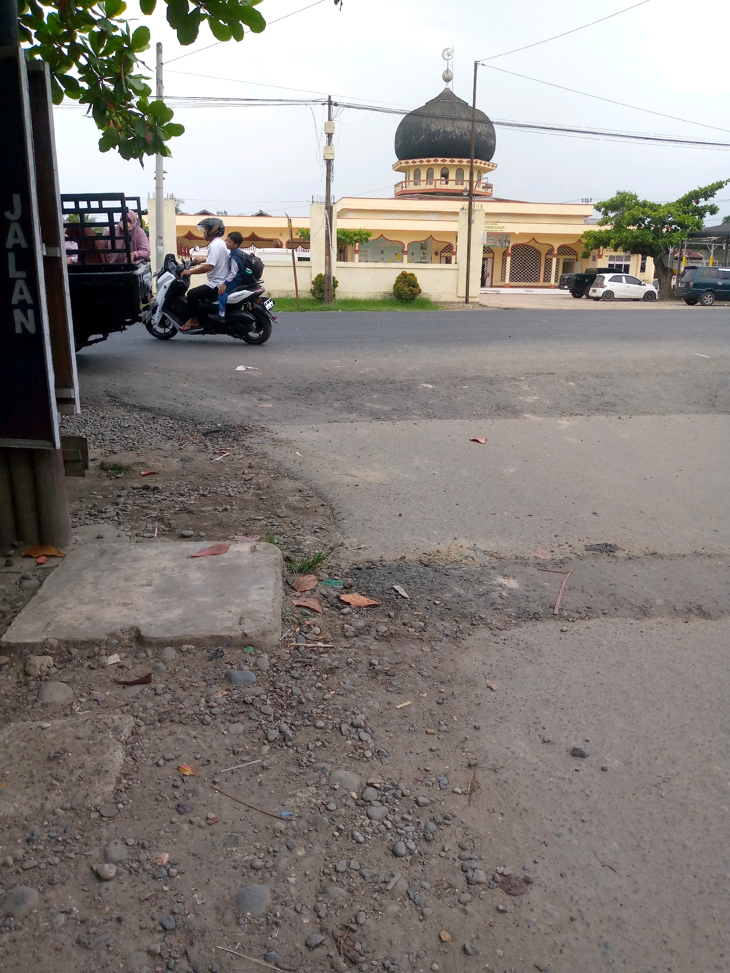 In Keudegeudong, Indonesia, a person on a scooter navigates a paved road. The scooter, a white and black Yamaha, carries a rider wearing a white shirt and helmet, and a child passenger in the back wearing a backpack. They are riding next to a black truck with a caged back, which is partially visible on the left side of the frame. The truck has "JALAN" written vertically on its side. In the background, a mosque with a prominent black dome and a light-colored building stands under a cloudy sky. Several cars are parked near the mosque, including a white car and a dark SUV. The road is lined with some vegetation and a utility pole. The ground is a mixture of pavement and loose gravel, with fallen leaves scattered around.