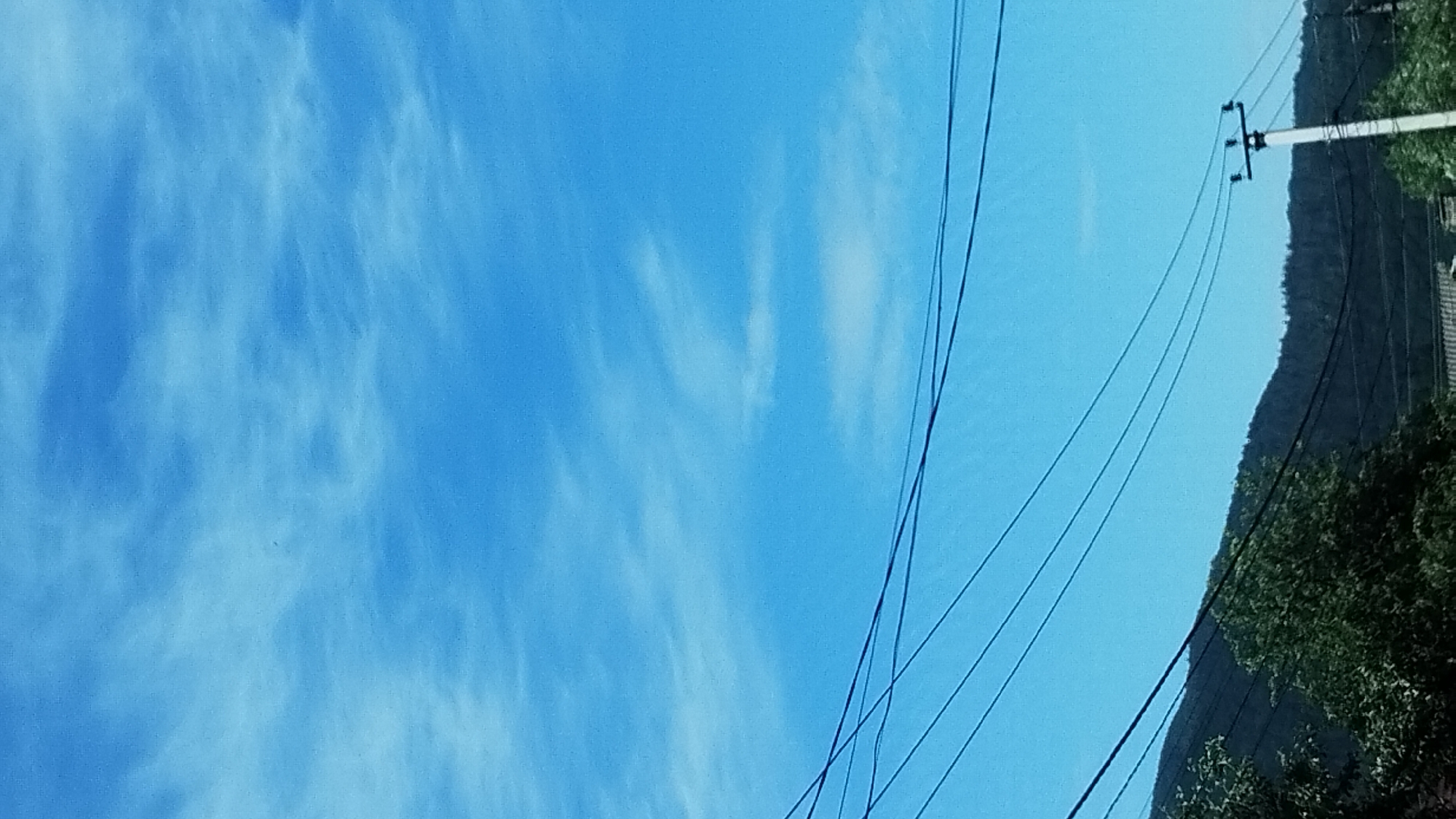 An outdoor, low-angle, vertical shot captures a bright blue sky with wispy, white clouds. Several dark power lines stretch horizontally across the frame, creating a layered effect against the sky. In the lower portion of the image, a dark, tree-covered mountain range is visible, with the silhouettes of trees in the foreground on the left. A white utility pole with crossbars and insulators stands on the right side of the frame, with additional power lines emanating from it. The scene appears to be an outdoor landscape with elements of both nature and infrastructure.