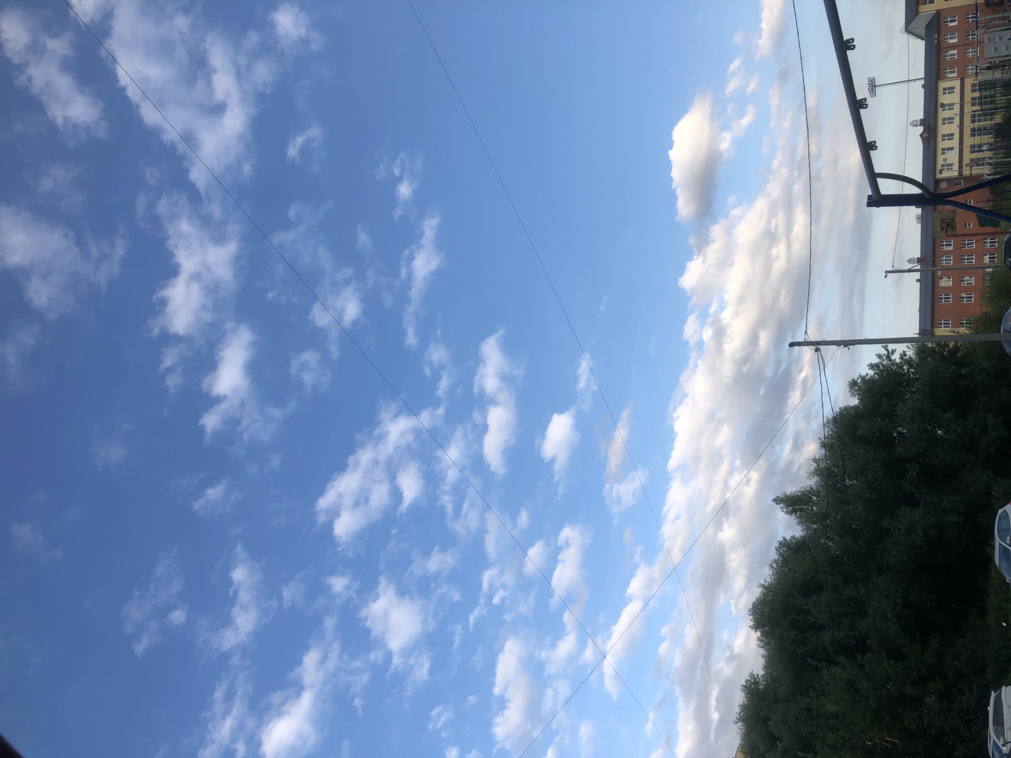 The image shows a blue sky with scattered white clouds. Several power lines crisscross the sky. In the lower portion of the image, a dense line of dark green trees is visible on the left, with the tops of buildings and a playground structure on the right. The buildings appear to be multi-story with numerous windows. A street light and a utility pole are also present. The location is identified as Rusanovo, Russia. There are no people visible in the image.
