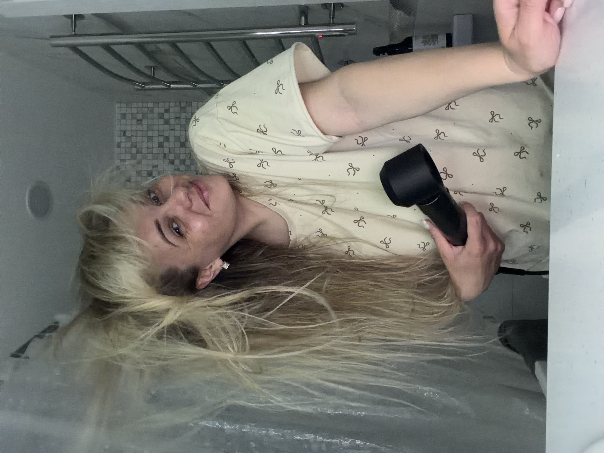 An indoor photograph, rotated 90 degrees counter-clockwise, features a person, likely female, with long, light blonde hair. The subject is shown from the chest up, with her head tilted towards the camera, displaying a faint smile. She is wearing a light-colored top patterned with small, dark, bow-like designs.
The person is actively engaged in holding a black hair styling tool, resembling a Dyson Airwrap, in her right hand. Her voluminous blonde hair is spread out, suggesting she is in the process of drying or styling it. Visible light-colored nail polish can be seen on her fingers.
The setting appears to be a bathroom, indicated by a metal towel rack mounted on a wall in the background. Part of the wall behind her head features small, gray, square tiles, while other areas are plain light-colored surfaces. A prominent white surface, possibly a counter or edge of a tub, frames the right side of the image. The lighting is consistent with an indoor environment. There is no legible text visible within the frame. This scene takes place in Votkinsk, Russia.