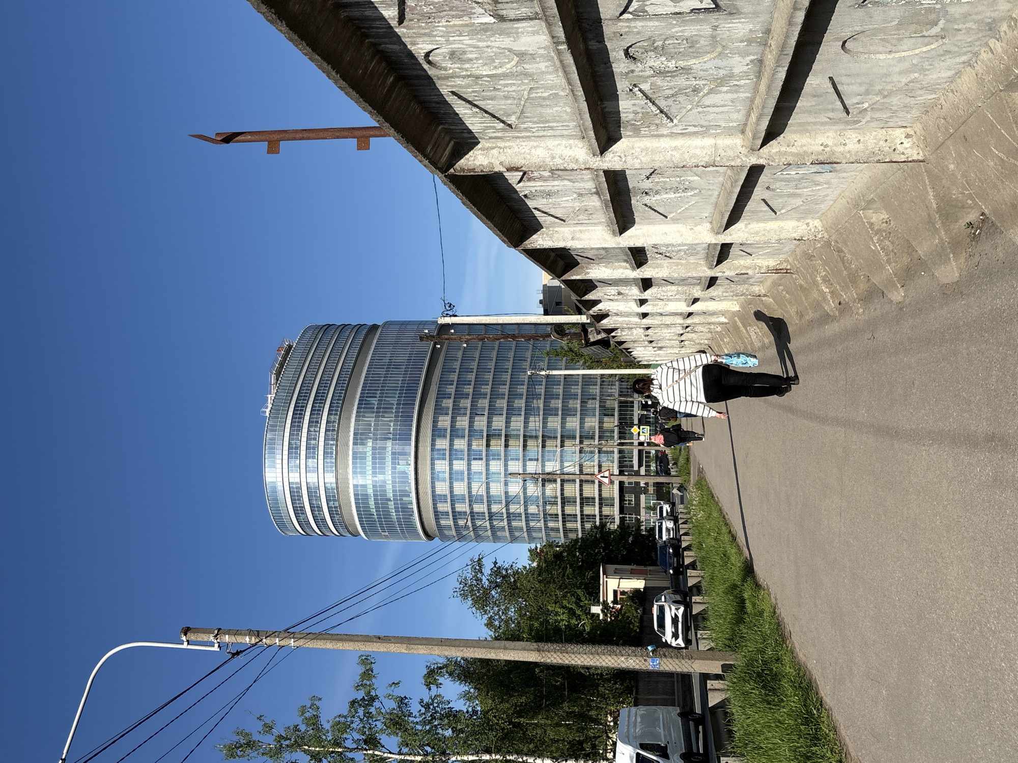 The image captures a bright, sunny daytime scene in an urban environment, viewed as if standing beneath a large concrete structure looking towards a modern building. A clear, vibrant blue sky dominates the upper left and central background.
In the foreground, a substantial concrete overpass or bridge structure spans the upper part of the frame. Its underside features geometric indentations and large, embossed letters that spell out "LOVE" clearly visible in sections. A rusted, L-shaped metallic bracket protrudes from the concrete towards the sky.
On the right side of the image, a paved pathway extends into the distance. A person, dressed in a white and black striped long-sleeved shirt and dark pants, is walking away from the viewer on this path, carrying a light blue bag. Their distinct shadow is cast long on the pavement, indicating strong sunlight.
To the left of the pathway is a strip of green grass, followed by a road where several cars are parked. Various trees and greenery are scattered along the roadside. In the lower left, a concrete utility pole with multiple power lines runs vertically.
Dominating the midground and left side of the scene is a tall, modern building with a curved or cylindrical design, featuring a facade made primarily of glass and metallic horizontal bands that reflect the blue sky. Near the base of this building, a red triangular warning sign and a blue square pedestrian crossing sign are visible on the roadside. The general setting suggests an area with both established infrastructure and contemporary development.