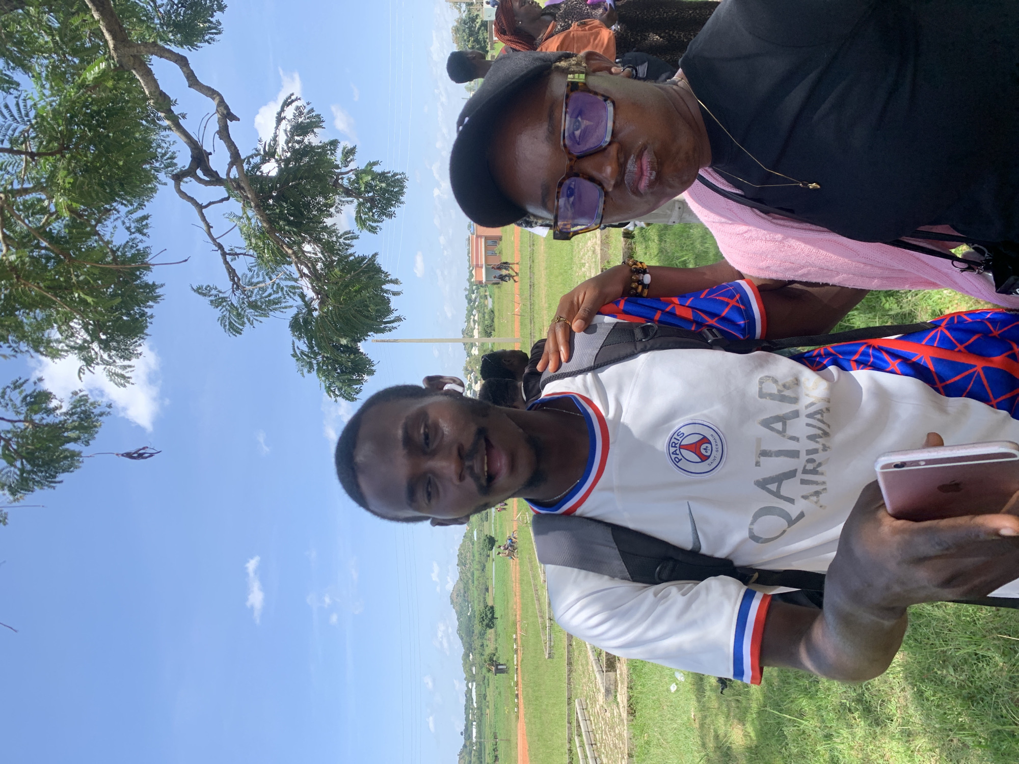 This is a vertical, selfie-style photograph taken outdoors in Gwafan, Nigeria. The primary subjects are two young Black men. The man in the foreground, on the right side of the image, is wearing a white Paris Saint-Germain soccer jersey with the Qatar Airways logo on it and blue and red trim on the sleeves. He is smiling at the camera and holding up a rose-gold colored smartphone in his right hand. He has a backpack on his shoulders.
Behind and to the left of the foreground subject is another young Black man wearing glasses and a black baseball cap. He is looking into the camera and appears to be speaking. He is also wearing a pink knitted garment underneath a black t-shirt, and a necklace.
In the background, there is a grassy field with some scattered trees. There are other people visible in the distance, some sitting and some standing, suggesting a gathering or event. A building with a red roof is visible in the far background. The sky is blue with a few scattered white clouds, indicating it is daytime and likely fair weather. The overall scene suggests a casual outdoor event or gathering.