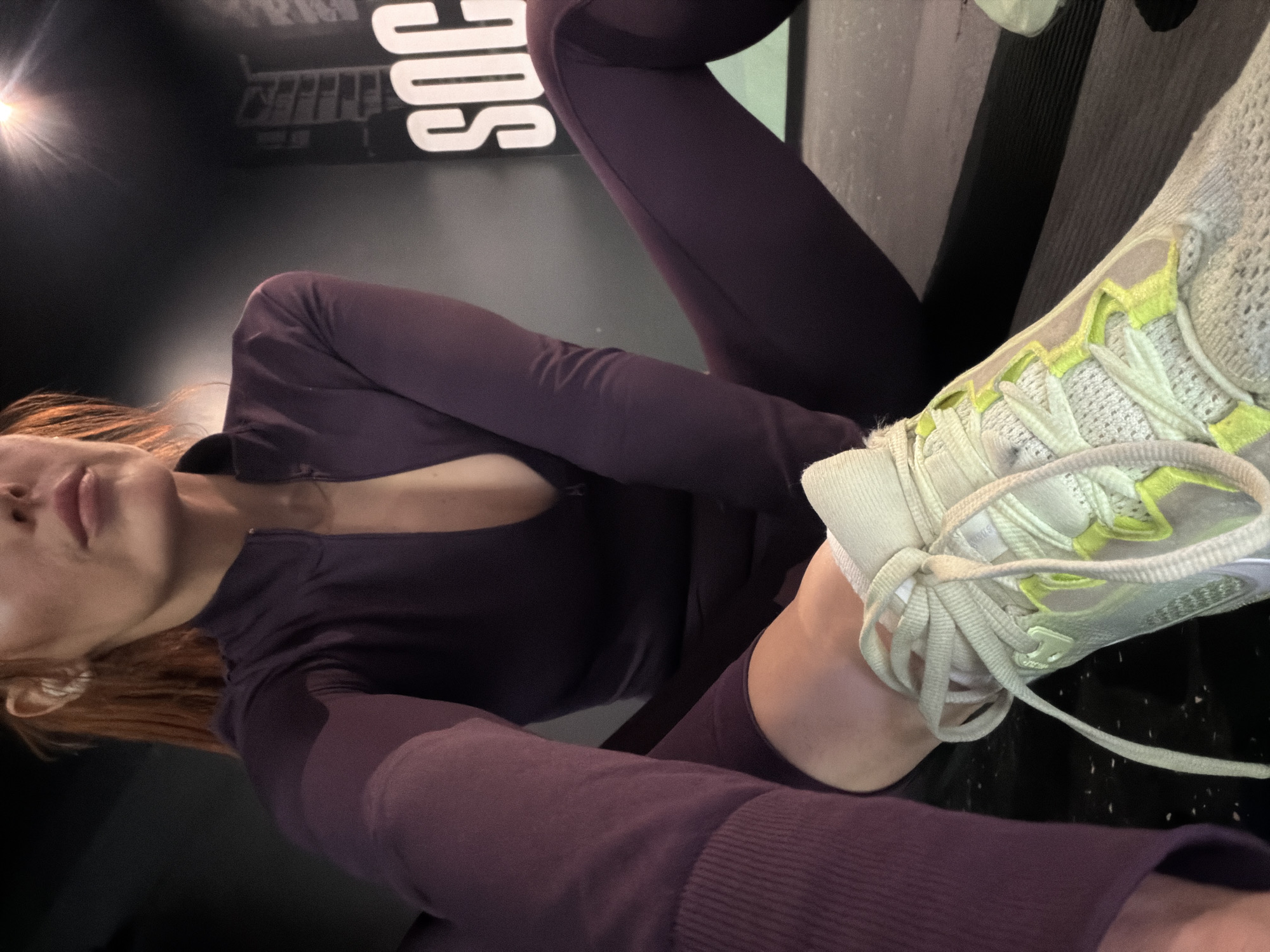 The image presents a close-up, low-angle shot of a person, likely female, wearing athletic attire and posing indoors. The individual has reddish-brown hair, and her face is partially visible from the chin to the lower nose, with lips appearing relaxed. She is dressed in a dark purple long-sleeved athletic top with a partial zipper, revealing some décolletage, and matching dark purple leggings.
One leg is lifted towards the camera, prominently displaying a light-colored athletic shoe, possibly off-white or very light grey, with distinctive neon yellow or lime green eyelets and light-colored laces. The shoe features a textured, mesh-like upper material.
The background consists of a dark, likely black, wall. Large white block letters spelling "SOC" are visible on the wall, and above them, faint rectangular outlines suggest further graphic elements or shelving. A bright, strong light source in the top-left corner creates a prominent lens flare, illuminating parts of the scene and contrasting with the otherwise dim environment.
The setting appears to be an indoor space, possibly a gym, fitness studio, or a modern leisure area, characterized by its dark decor and artificial lighting. The person seems to be casually posing for a photograph. The external context indicates the location is Paris, France.