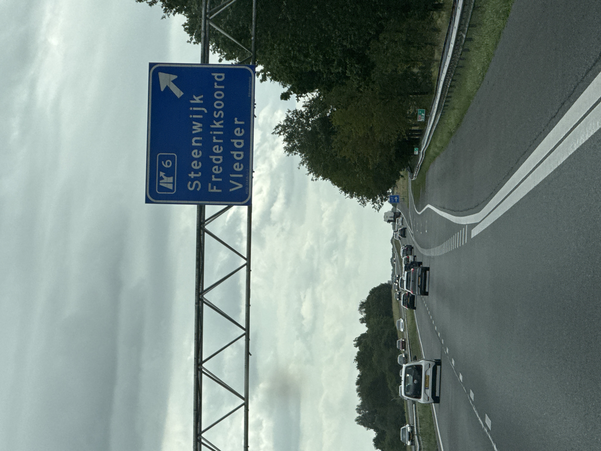 The image depicts a highway scene on a cloudy day. A blue overhead road sign displays directions for "Steenwijk," "Frederiksoord," and "Vledder," with an arrow indicating the direction for these locations. The sign also shows "A6" and a smaller symbol. Numerous cars are driving on the multi-lane highway. To the right of the highway, there is a guardrail and dense green trees. On the left side of the highway, there are more trees and a slightly elevated embankment. The road appears to be in or near the city of Tuk, Netherlands, given the destination names on the sign. The weather is overcast, with a gray sky.