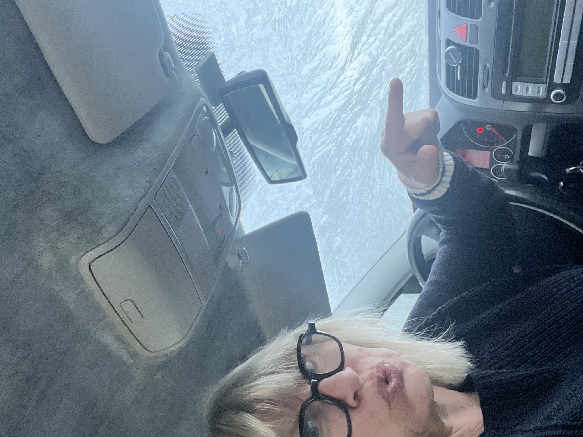 The image, taken from inside a vehicle, shows a person with blonde hair and glasses giving the middle finger. The vehicle's dashboard and part of the steering wheel are visible, along with the rearview mirror and sun visor. The windshield is covered with what appears to be ice or frost. The location is not discernible as Chambœuf, France.