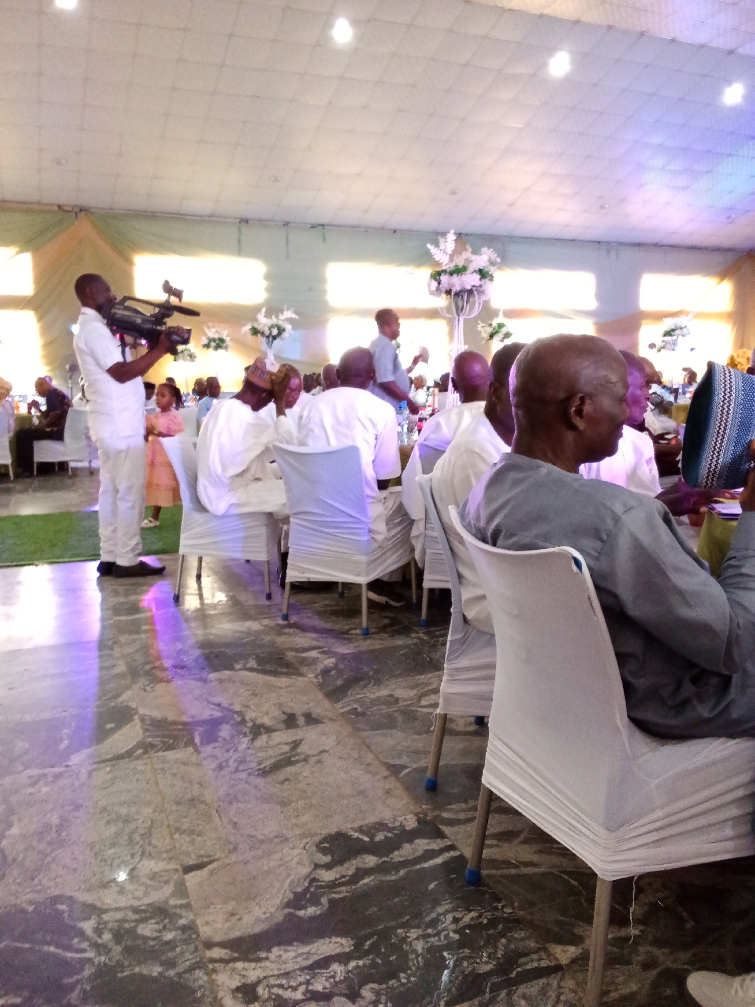 In an indoor venue, likely a reception hall, people are seated at tables covered with white cloths, with some chairs also draped in white. A man in a white shirt and pants operates a video camera. A young girl in a pink dress stands nearby. The floor is tiled with a marbled pattern. Floral arrangements are visible on pedestals. The ceiling is paneled, with lights illuminating the space, and colored lights cast reflections on the floor. A man in a grey garment is seated and holding a blue and white patterned hat. The scene appears to be a formal gathering or event.