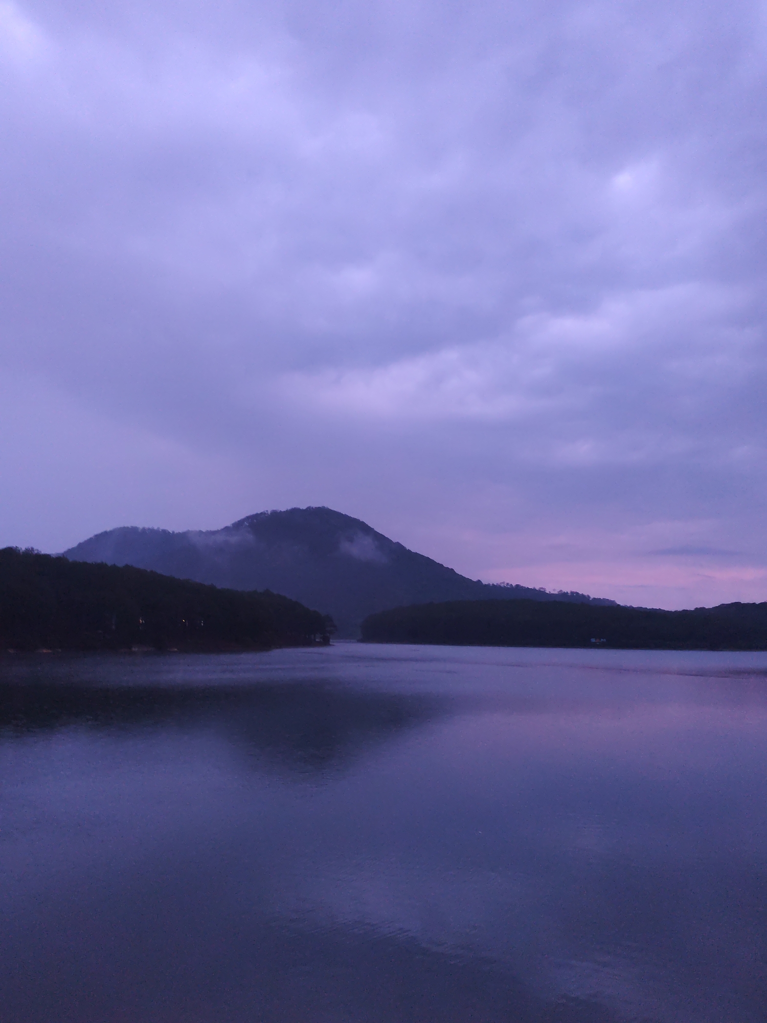 The image depicts a twilight or pre-dawn scene on a body of water, likely a lake, surrounded by hills or mountains. The sky is overcast with hues of purple and gray, suggesting a time shortly before sunrise or after sunset. A larger, prominent hill is partially shrouded in mist or low clouds. The water's surface reflects the colors of the sky and the dark silhouettes of the surrounding landmasses. There are no visible people or distinct objects other than the landscape itself. The scene conveys a sense of stillness and tranquility. There is nothing to indicate the location is Da Lat, Vietnam, besides general features such as a lake and low-lying mountains.