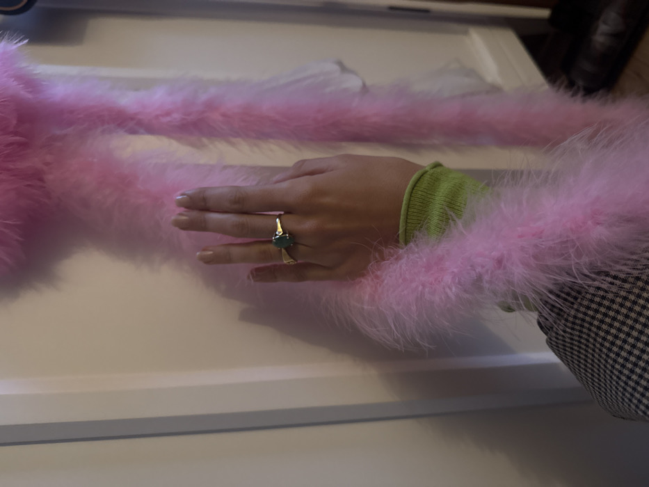 The image presents a close-up, top-down view of a person's hand and arm, adorned with various accessories and clothing items. The arm extends from the right side of the frame towards the left.
The hand is positioned with fingers slightly spread and relaxed, palms down. A prominent gold-toned ring with an oval-shaped, dark green or teal gemstone is worn on the finger, possibly the middle or ring finger, on the left hand. The fingernails are neatly manicured with a neutral or light polish.
The arm is clothed in a light, almost lime-green long-sleeved garment, possibly a sweater or a top, with a distinct ribbed cuff visible at the wrist. This green sleeve is partially obscured by a voluminous, fluffy pink feather boa or trim that surrounds the arm and extends across the width of the image. The feathers are soft and appear to be part of an elaborate cuff or a separate accessory draped around the arm. On the far right, a small portion of a patterned fabric, resembling a black and white houndstooth or gingham check, is visible, suggesting another layer of clothing, perhaps a jacket sleeve.
The background is a smooth, light-colored surface, likely a table or countertop, possibly white or off-white, which provides a clean contrast to the vibrant pink feathers and green sleeve. Shadows are cast on this surface, particularly beneath the hand and feathers, indicating a light source from above or the left.
The overall impression is one of fashion, style, or personal adornment, with a focus on contrasting textures and colors: the smooth skin, the ribbed green fabric, the soft fluffy pink feathers, and the checkered pattern.