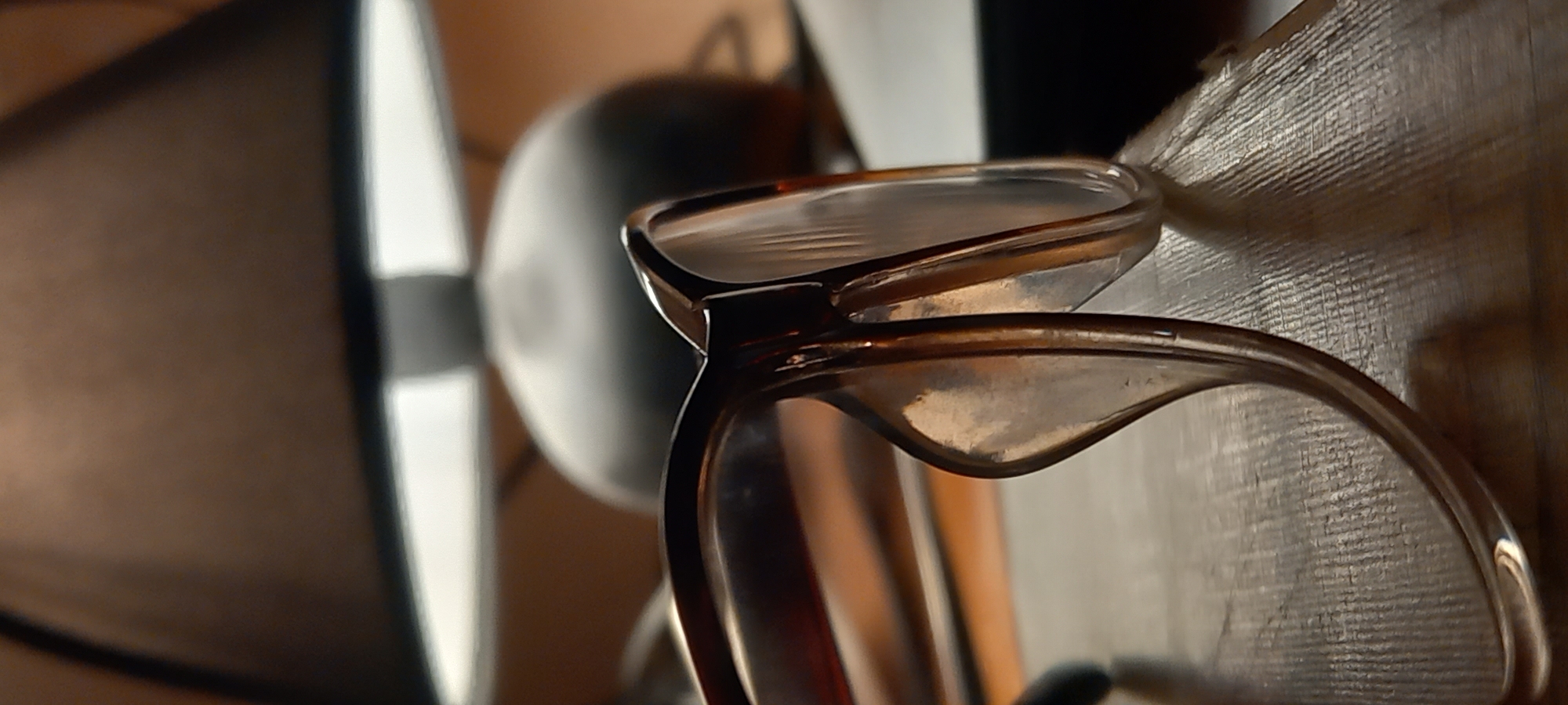 The image presents a close-up, shallow-depth-of-field shot of a pair of eyeglasses, likely taken indoors in Kryvyi Rih, Ukraine. The main subject is a pair of brown or amber-colored eyeglasses with clear lenses, positioned diagonally and in sharp focus in the mid-ground. The frames appear to be made of plastic or acetate, with warm reflections visible on their surfaces and lenses.
The background is heavily blurred, creating a soft, out-of-focus effect that highlights the eyeglasses. To the far left, a large, dark, indistinct object with a bright vertical edge is visible, possibly a lamp shade, a fan blade, or part of a speaker. Directly behind the eyeglasses, another large, dark, rounded shape suggests a piece of equipment or furniture. To the right of the eyeglasses, there is a textured, light-colored surface, possibly a wooden wall, woven material, or a piece of textured furniture, displaying fine horizontal lines. The lighting is warm, suggesting indoor illumination. There is no discernible activity taking place, and no people are visible. No text is present in the image. The overall impression is a quiet, still-life composition of everyday objects in a domestic setting.