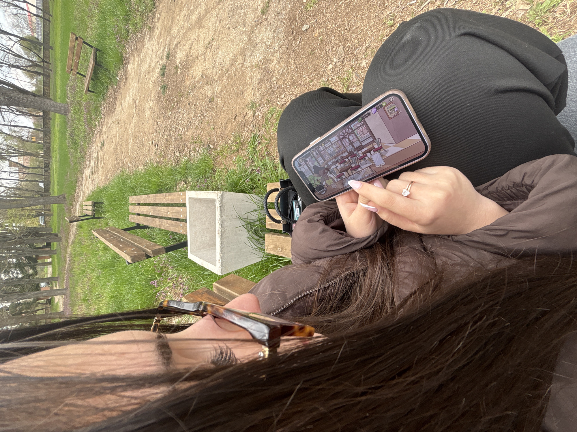A woman with long, dark brown hair and glasses is sitting on a park bench in what appears to be a park or green space. She is wearing a brown jacket and black pants. She is holding a smartphone and looking at the screen, which displays a cartoon-style interior scene. The woman's hand has pale pink nail polish and a ring. In the background, there is a dirt path, patches of green grass, trees, and other park benches. One bench has a concrete cube structure attached to it. Based on the location information, this scene is located in Lehchevo, Bulgaria.