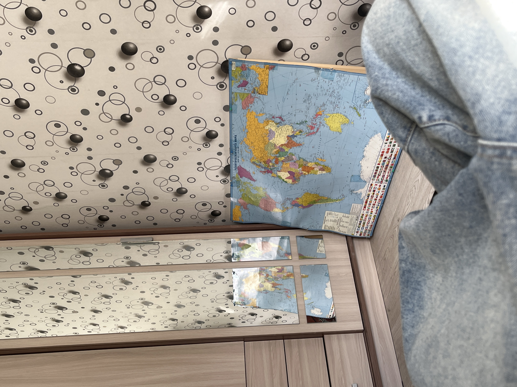 A high-angle indoor shot shows a world map leaning against a wall with patterned wallpaper above a dresser. The dresser has a mirrored top, reflecting the map and wallpaper. The right side of the frame is partially occupied by a large, light-blue denim jacket or shirt. The dresser appears to be made of light-colored wood with a grain pattern. The wallpaper is light with a pattern of black dots and gray circles. The location is Unknown, Unknown. No people are visible.