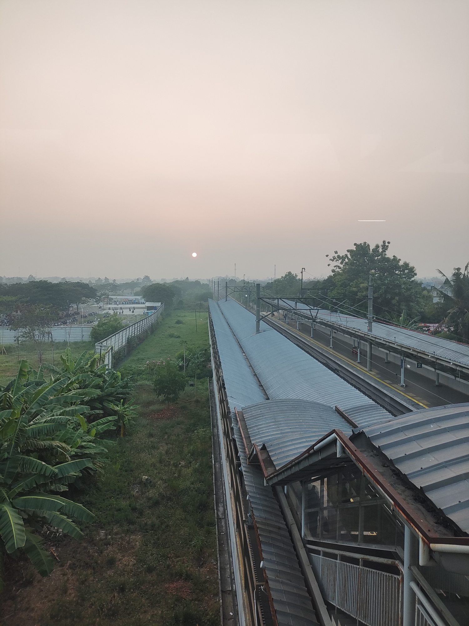 The image shows a railway station in Telagamurni, Indonesia, at sunrise. The sky is hazy and a pale orange hue, with the sun appearing as a small, bright circle in the upper left quadrant of the image. In the foreground, there is a covered walkway or bridge over the train tracks, with a corrugated metal roof and a rusted metal railing. To the left of the tracks, there is a grassy area with banana trees and other vegetation. Beyond that, there is a fence, a road with parked vehicles, and more trees in the distance. To the right of the tracks, there is another covered platform and more trees. The scene appears to be quiet and tranquil, with no people visible on the platforms or tracks. The overall impression is one of a typical morning scene at a suburban train station.