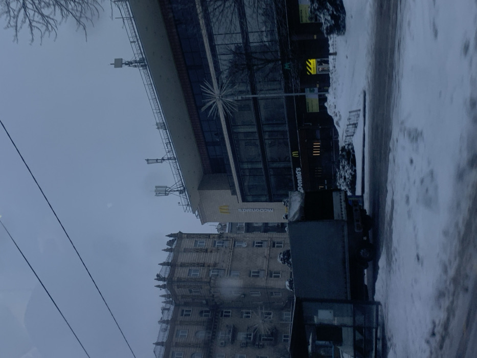 The image depicts a winter street scene on Khreshchatyk Street, 19A, in Kyiv, Ukraine, during overcast daylight. The ground, rooftops, and vegetation are covered in a layer of white snow.
In the mid-ground, a multi-story modern building with a dark, reflective glass facade prominently features a McDonald's restaurant. A large vertical "McDonald's" sign with the golden arches logo is visible on the upper left side of the facade, and another horizontal "McDonald's" sign is at street level. The building's flat roof is equipped with multiple telecommunication antennae. In front of this building, a large, white, star-shaped decorative fixture is mounted on a pole. To the left of the McDonald's building, an older, multi-story architectural structure with classical details, stone construction, and balconies stands.
In the foreground, a light-colored cargo truck or van with a dark grey tarp-covered bed is partially visible on the left, positioned near what appears to be a bus stop shelter. The street in front is snow-covered, with darker sections indicating cleared or melted areas. Overhead wires, likely for trolleybuses, traverse the upper portion of the frame against the grey sky. Snow-covered bushes and a black metal fence are visible along the sidewalk area. No people are discernible in the frame.