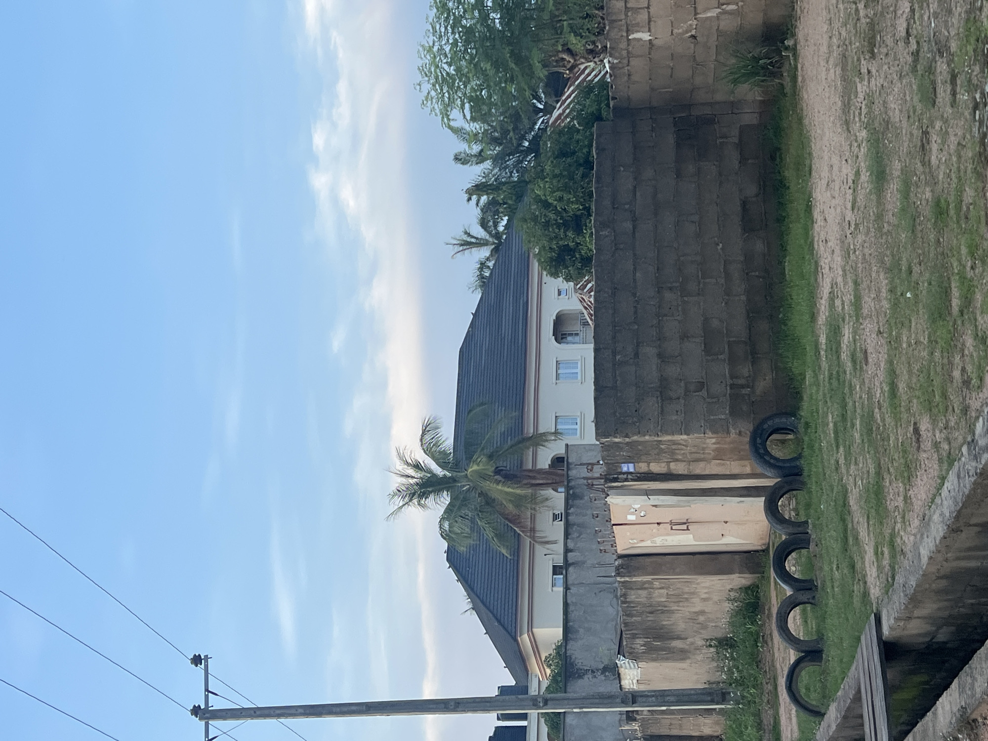 The image depicts a residential area in Jalingo, Nigeria, under a cloudy blue sky. A light pole with multiple wires stands on the left side of the frame. In the background, a large, modern house with a dark tiled roof is visible behind a concrete block wall and a chain-link fence. A prominent palm tree grows in front of the house. Several smaller trees and greenery are scattered throughout the scene. In the foreground, a low concrete wall runs along the bottom of the image, with a small wooden plank bridge crossing over a drain. Several car tires are arranged on the grassy ground in front of the wall. The overall impression is of a typical urban or suburban environment in Nigeria.