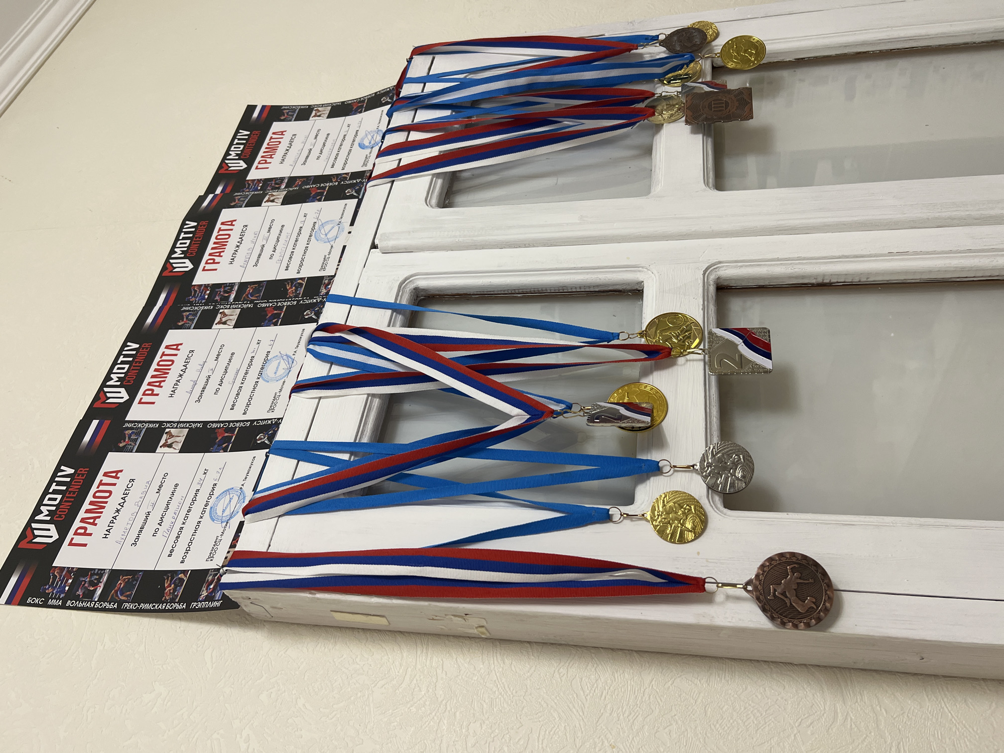 The image displays a close-up view of several medals hanging from a white wooden surface, likely a door or cabinet. Above the medals, three certificates with the "MOTIV CONTENDER" logo are affixed to the wall. The certificates are in Russian and appear to be awards for athletic achievements, with fields for the winner's name, ranking, discipline, weight category, and age category. The medals themselves are of various metals, including gold, silver, and bronze, and feature engraved designs and red, white, and blue ribbons. The environment appears to be indoors, with a textured wall in the background. The certificates and medals suggest a connection to sports or martial arts competitions. The specific location of Sochi, Russia, is not evident from the visual information provided in the image itself.