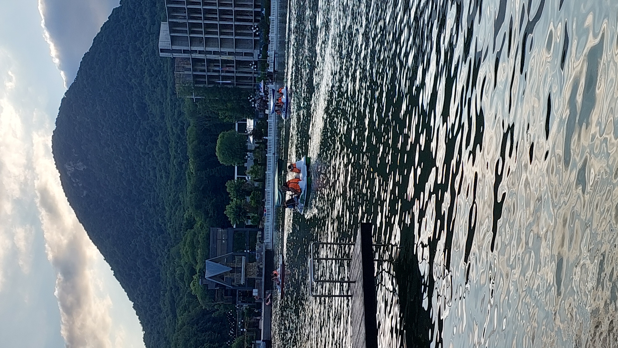 This is a photograph taken at the ZHELEZNOVODSK city park in Zheleznovodsk, Russia. The image depicts a lake with several paddle boats carrying people. In the background, a large, tree-covered mountain dominates the skyline, with a partly cloudy sky above. To the right of the mountain, a multi-story building, likely under construction, is visible. On the left side of the lake, there is a wooden dock and a modern-looking building with a distinctive roof structure. The water in the lake is rippled, reflecting the sky and surrounding landscape. The overall scene suggests a recreational area on a partly cloudy day.