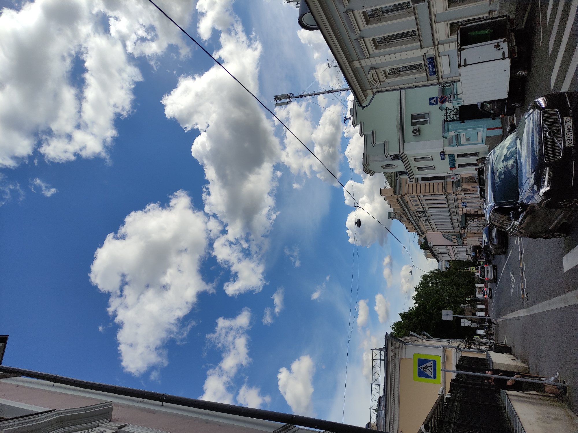 The image depicts a street scene in Moscow, Russia, under a partly cloudy blue sky. The road is lined with buildings, predominantly in light colors, some of which feature ornate architectural details. Various vehicles, including a black Volvo SUV and a white delivery truck, are parked or moving along the street. A pedestrian crossing sign is visible, and a person is leaning against the metal railing. Overhead power lines stretch across the sky.