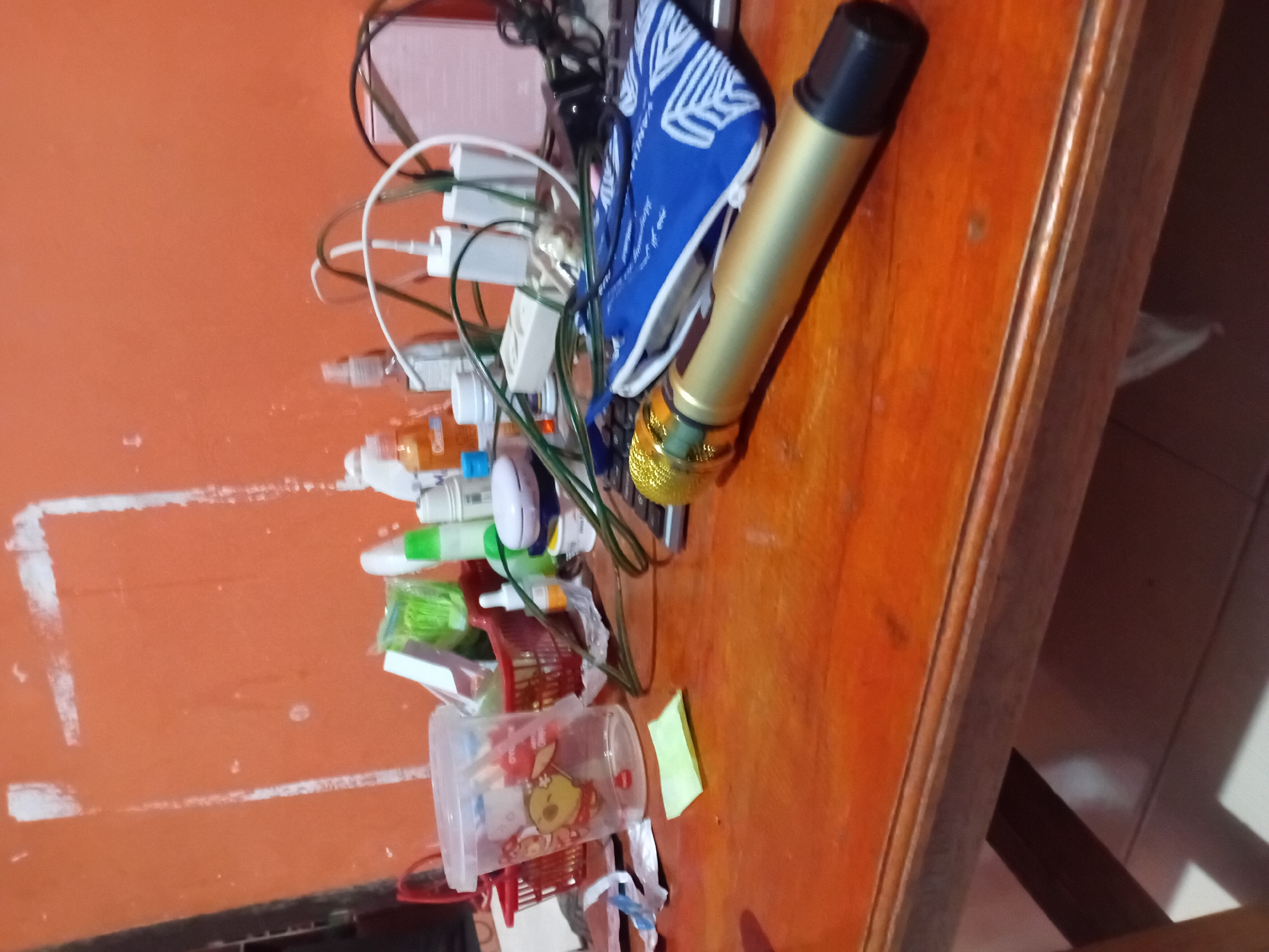The image displays a wooden desk in Kasang Pudak, Indonesia, cluttered with various items. Prominently featured is a gold and black microphone lying horizontally across the desk. To the left of the microphone, a blue pouch with white leafy patterns is visible, partially covering a black keyboard. In the background, a collection of toiletries and electronic accessories, including charging adapters and cables, are scattered on an orange surface, possibly a shelf or wall. A clear plastic cup with cartoon decals and a small red basket are also present on the desk. The overall scene suggests a personal workspace with items for entertainment and daily use.