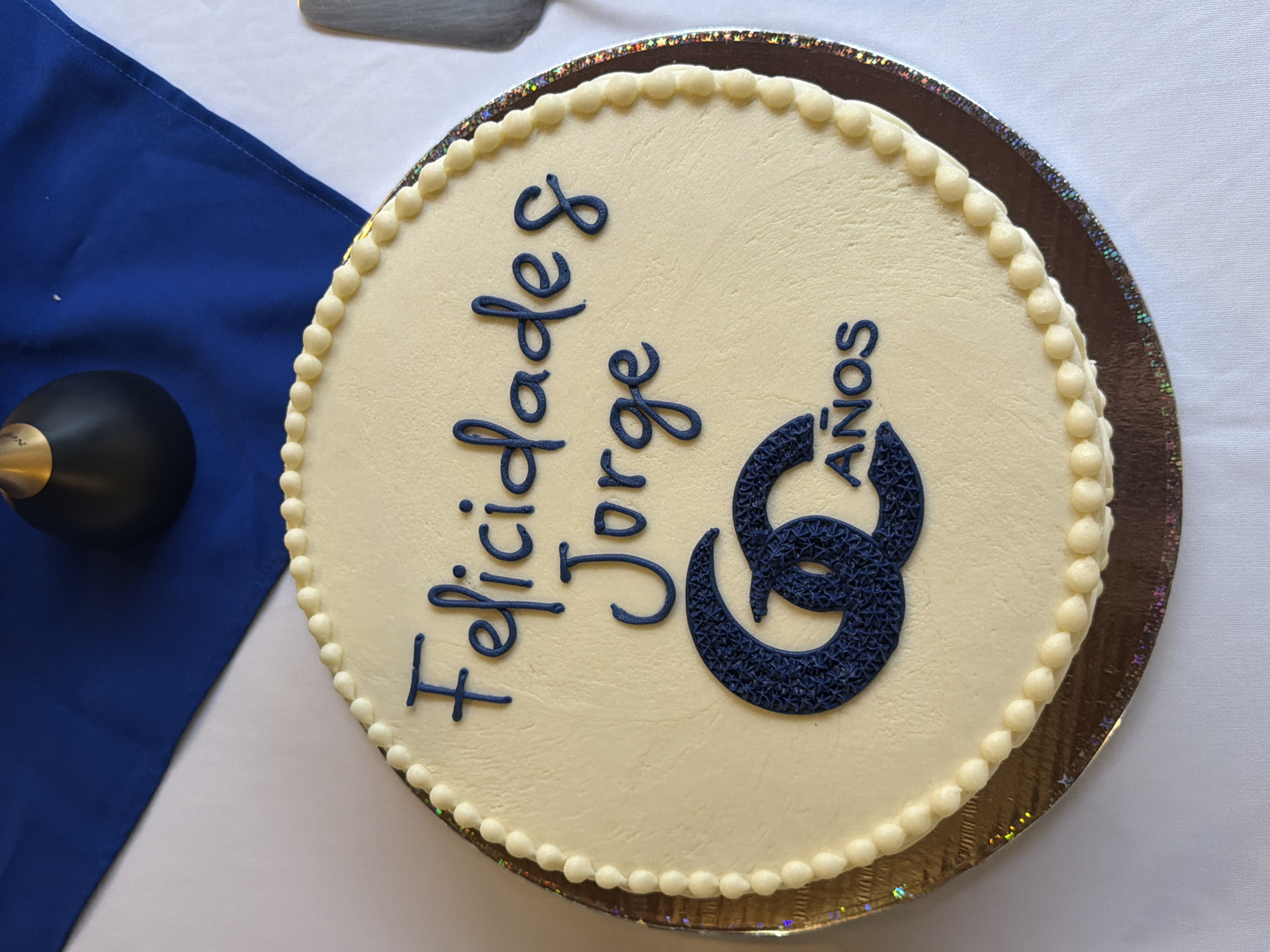 The image shows a round cake with white frosting, decorated with dark blue lettering that reads "Felicidades Jorge 60 Años." The cake is on a gold-colored cardboard cake board. To the left of the cake, a portion of a blue cloth napkin is visible, along with a black object with a gold accent. The cake and napkin appear to be on a white table. The location is Unknown, Unknown.