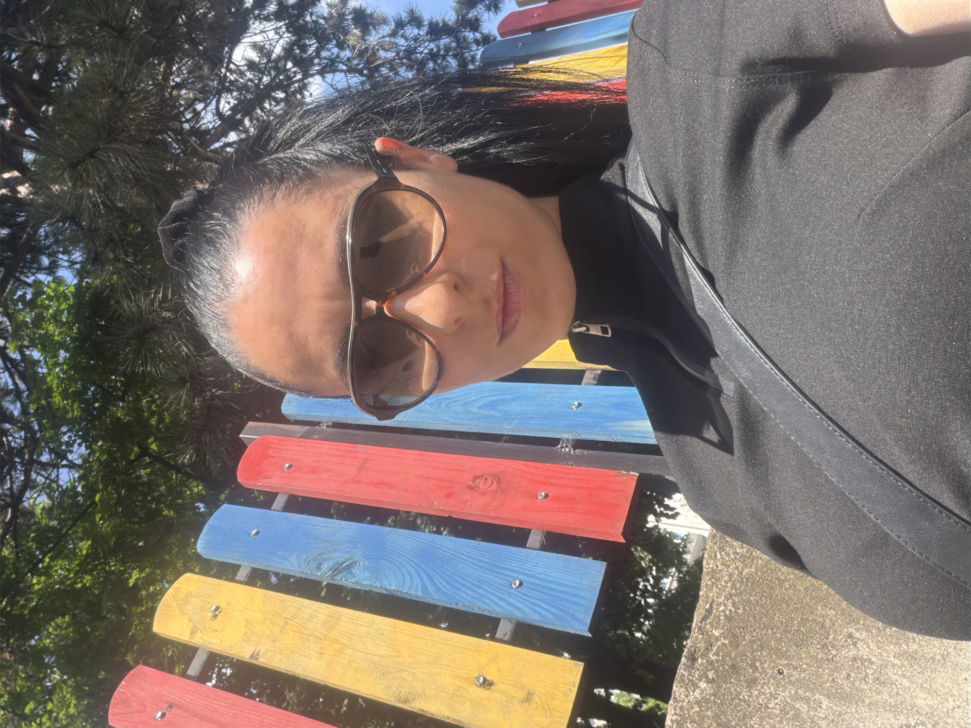 A woman with dark hair pulled back in a bun and wearing sunglasses is sitting on a vividly painted wooden bench outdoors. The bench has red, blue, and yellow planks. She is wearing a black jacket and has a black strap over her shoulder. Trees are visible in the background under a blue sky. The location is likely a park in the city of Pleven, Bulgaria.