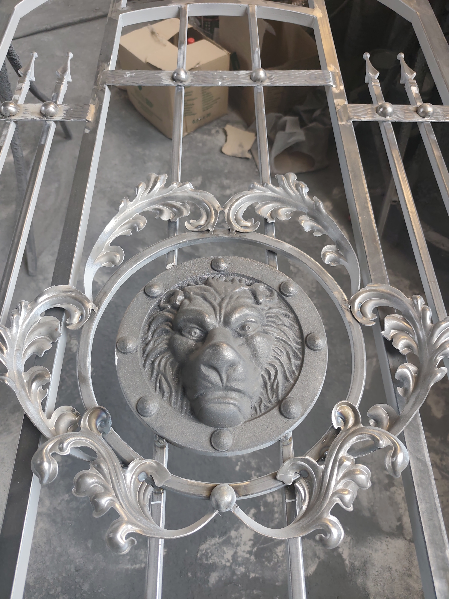A close-up, eye-level shot captures a partially constructed metal gate, likely an entryway, with ornate detailing. The gate is composed of vertical metal bars, some topped with pointed finials, and horizontal crossbars. A prominent circular medallion featuring a relief of a lion's head, detailed with a mane and fierce expression, is centered within the gate. Surrounding the medallion are decorative metal scrolls and leaf-like embellishments, also in a silver-toned metal. The background consists of a rough concrete floor and a partially visible cardboard box, suggesting an unfinished construction or workshop environment. The lighting is even, with no strong shadows, indicating an indoor setting.