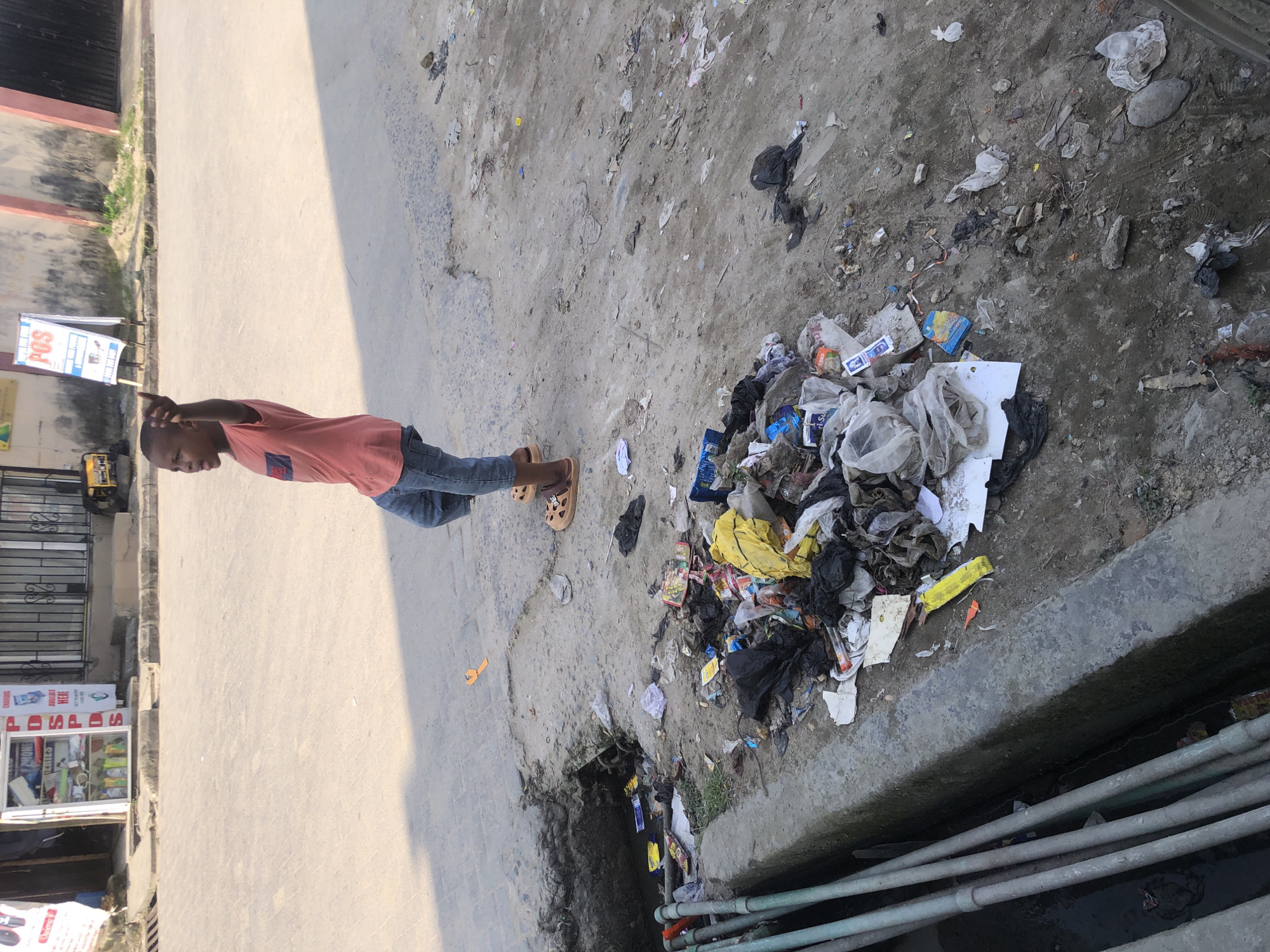 A young boy stands in the middle of a dusty road, raising his right hand as if gesturing or pointing. He is wearing a coral-colored t-shirt and denim shorts, with brown sandals on his feet. The ground around him is littered with trash, including plastic bags, wrappers, and other debris. A drainage ditch filled with murky water and more debris is visible to the left. In the background, there are buildings and a generator, suggesting an urban setting, possibly in Ado, Nigeria. The sun casts long shadows across the scene.