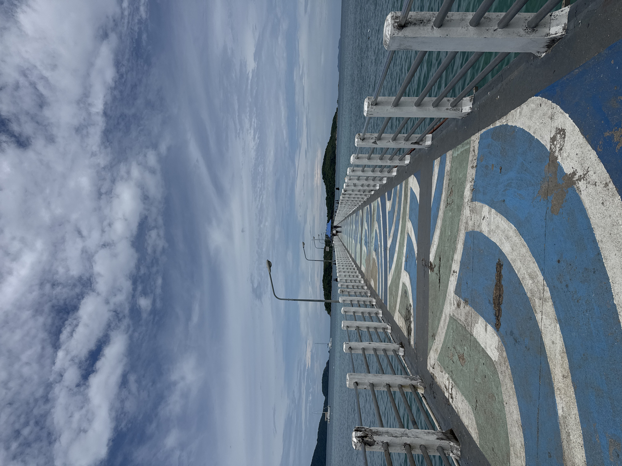 The image depicts a pier extending into the sea in Rawai, Thailand. The pier's surface is painted with a repeating pattern of blue, white, and green curved lines. A white metal railing with concrete posts runs along both sides of the pier. Several streetlights are visible along the pier, spaced at regular intervals. In the distance, islands covered in lush green vegetation can be seen. The sea is a deep blue-green, with small waves. Two sailboats are anchored offshore. The sky is partly cloudy, with dramatic streaks of white clouds against a blue background. A few people are walking on the pier in the distance, appearing as small figures against the vastness of the sea and sky. The scene evokes a sense of calm and openness.