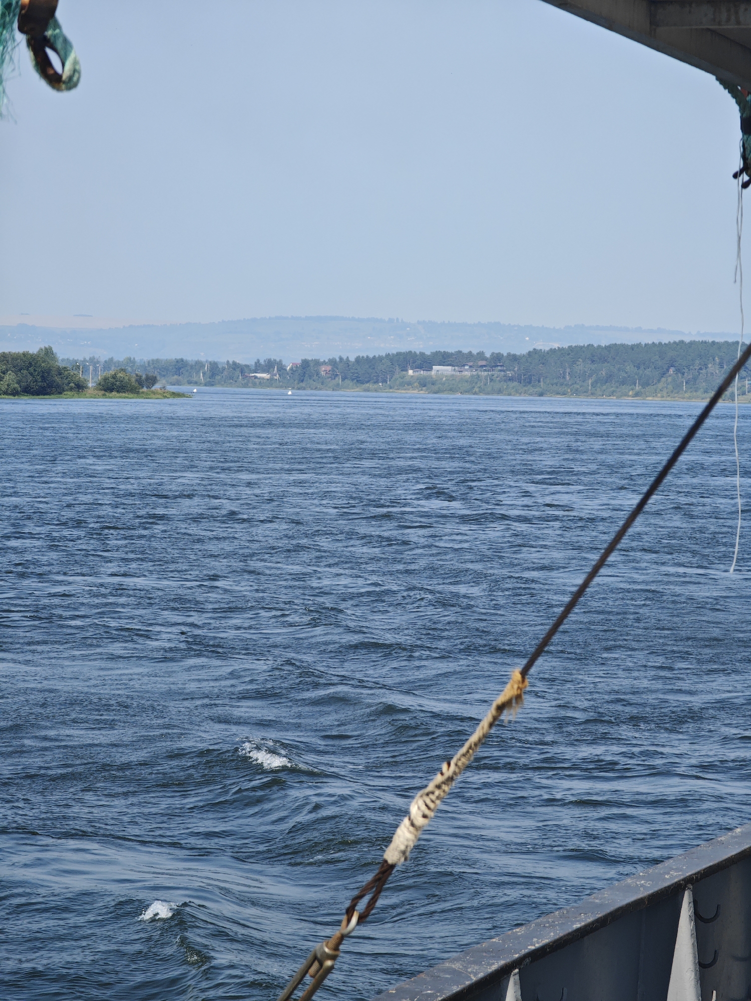 The image captures a wide, choppy blue river under a clear, pale sky. The foreground is dominated by the metal railing of a vessel, with a thick cable extending diagonally across the frame, wrapped partially with a patterned cloth. A green net and a metal ring are visible hanging from the upper left corner of the image, also attached to the vessel. In the middle ground, a tree-lined bank forms the horizon, with scattered buildings and structures visible amongst the foliage. The river is the primary subject, showing ripples and waves across its surface, suggesting movement and possibly a moderate current. The overall impression is one of a journey or a view from a boat on a river, likely during daylight hours. The scene is devoid of human figures, focusing instead on the natural and man-made elements of the environment.
While the specific location of Krasnoyarsk, Russia, is provided as context, the visual information within the image itself does not offer any direct clues to pinpoint this exact geographical area. The landscape is generic to many riverine environments and lacks distinctive landmarks or architectural styles that would definitively tie it to Krasnoyarsk.