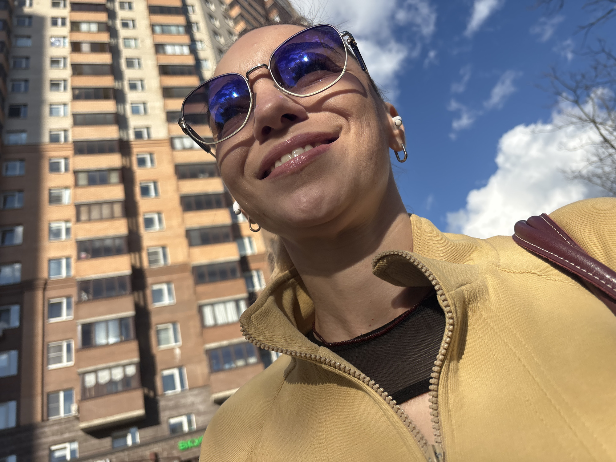 A low-angle, close-up shot captures a smiling woman in sunglasses, looking upwards. She has blonde hair pulled back and is wearing a yellow zip-up jacket over a black top, with a thin beaded necklace. In the background, a tall, beige apartment building with many windows dominates the left side of the frame, under a bright blue sky with scattered white clouds. A hint of bare tree branches is visible on the right. The scene appears to be an outdoor urban setting, likely in Russia, given the architectural style of the building.