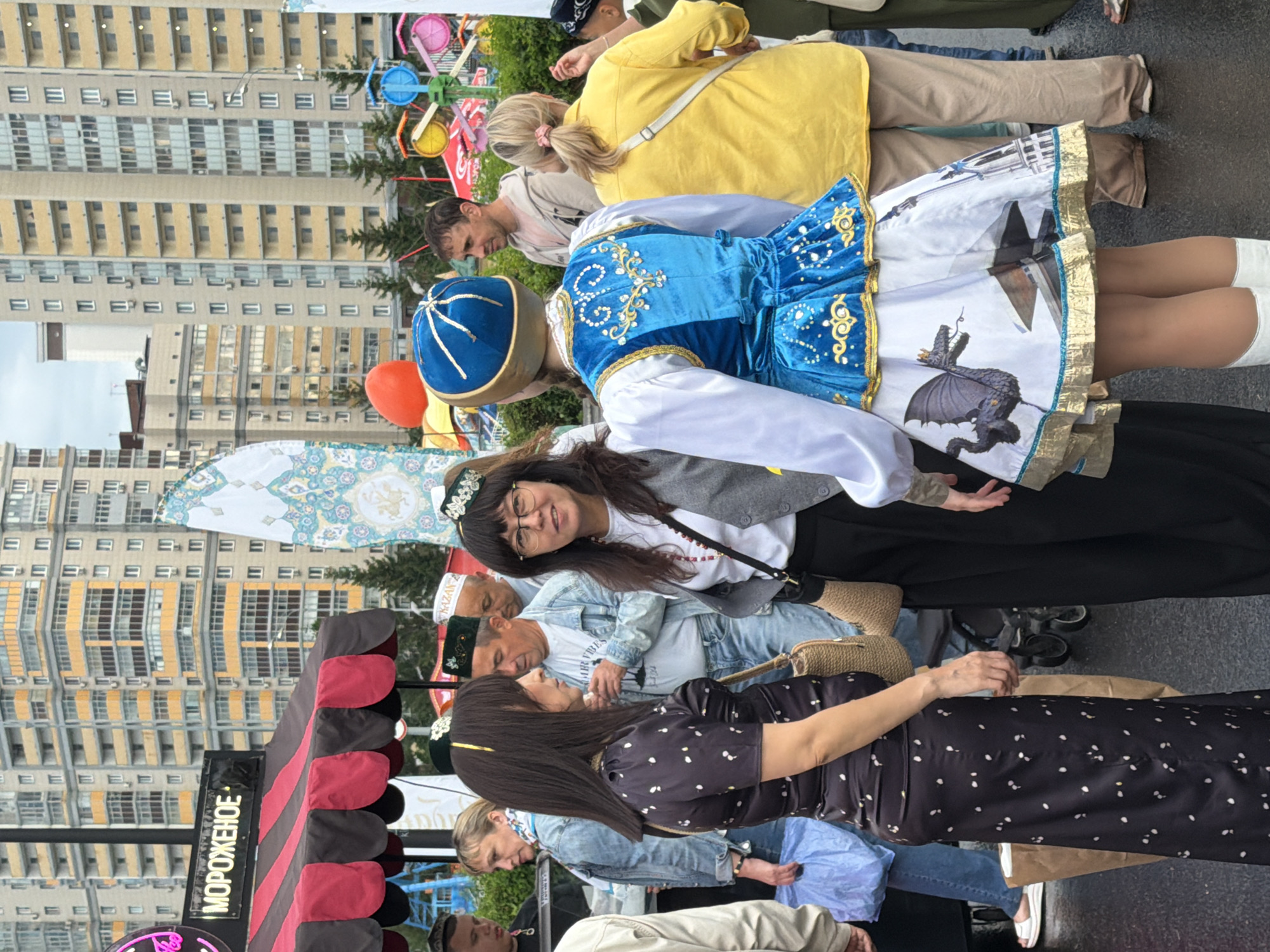 In the city of Unknown, Unknown, a group of people are gathered outdoors. A woman in a long, dark, polka-dotted dress stands in the foreground, facing left. Next to her, a woman wearing glasses and a gray vest over a white shirt looks towards the camera. To the right of the woman in the vest, a person in a vibrant blue and gold traditional outfit, with a skirt featuring a dragon and building print, faces away from the camera. Behind them, several other individuals are visible, including a man in a light-colored hoodie and a woman in a bright yellow shirt. In the background, tall residential buildings with yellow and gray facades dominate the skyline. A red and black striped awning of what appears to be an ice cream stand is visible on the left, with text in Russian reading "МОРОЖЕНОЕ" (ice cream). Various decorative flags and a colorful amusement ride are also present in the scene. The ground appears wet, suggesting recent rain.