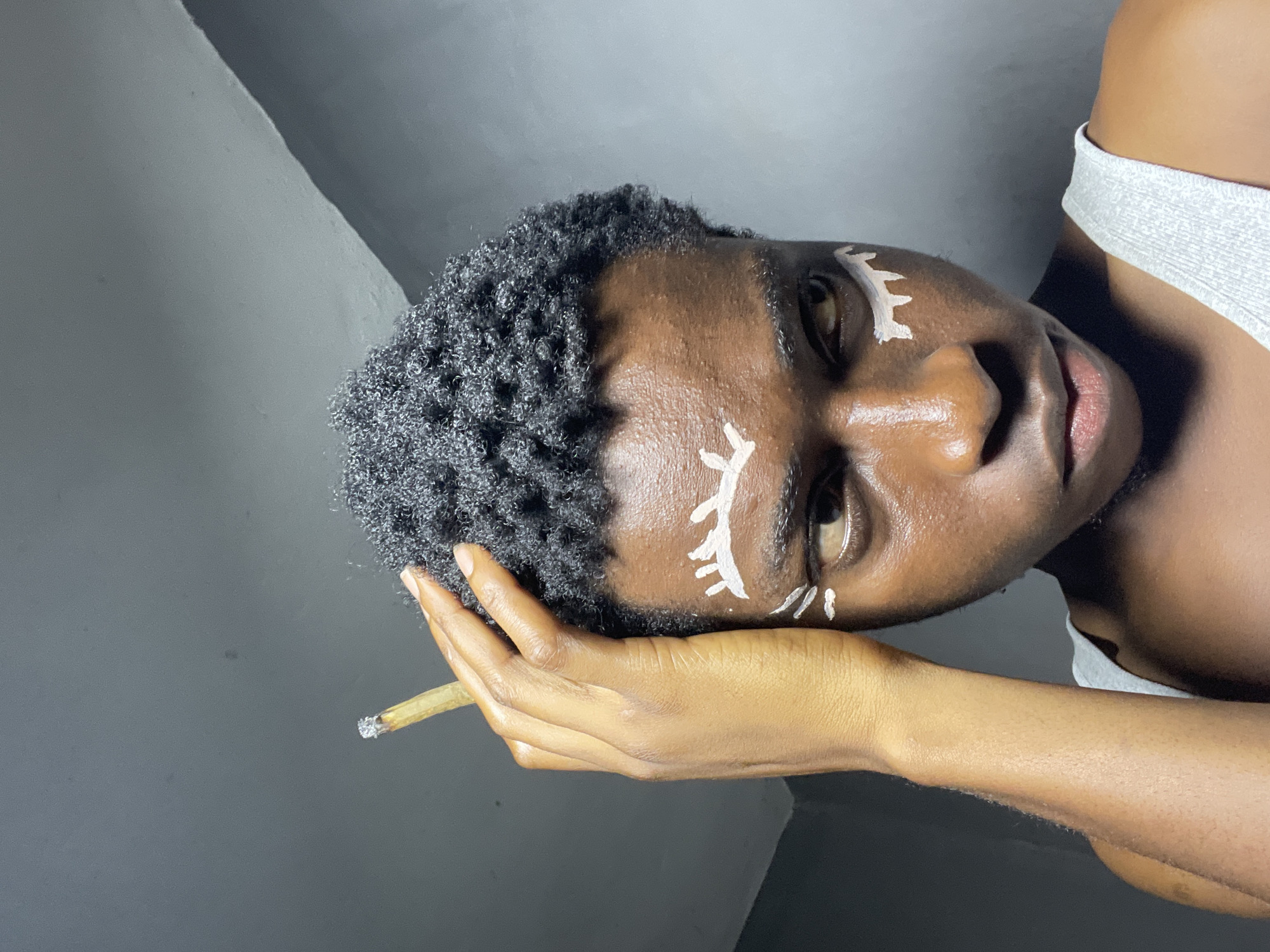 A close-up, waist-up shot shows a young Black person with short, dark, curly hair and dark skin, wearing a light grey tank top. White face paint is applied in the style of stylized eyelashes above and below each eye, and also as dashes below the painted eyelashes on the right cheek. The person is looking to their right, with their left hand raised to touch their hair, holding a lit cigarette. The background is a plain, grey wall. This image was captured in Nkpor, Nigeria.