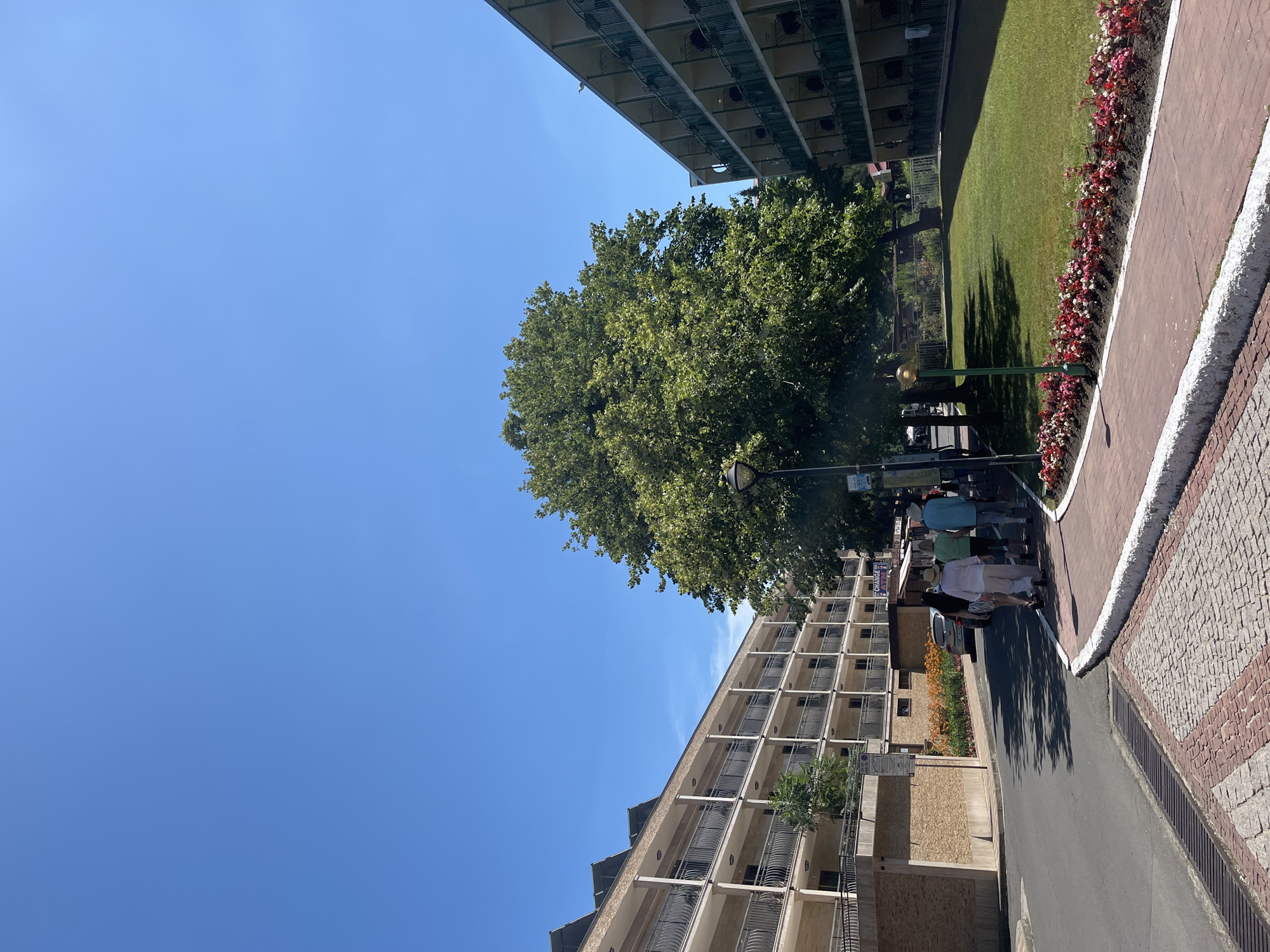 A group of people are walking along a sidewalk next to a street. The scene is set in Nesebar, Bulgaria, on a sunny day. Tall apartment buildings with balconies line one side of the street, while a grassy area with a flowerbed separates the sidewalk from a building on the other side. The sky is a clear blue, suggesting it is daytime. The people are dressed in casual summer clothing. The time of day appears to be late morning or early afternoon, given the angle of the shadows. The atmosphere seems relaxed and pleasant, typical of a warm day in a tourist area. The architecture of the buildings and the paving stones of the sidewalk provide some context for the location. There is some signage visible on the buildings, but the text is not legible.