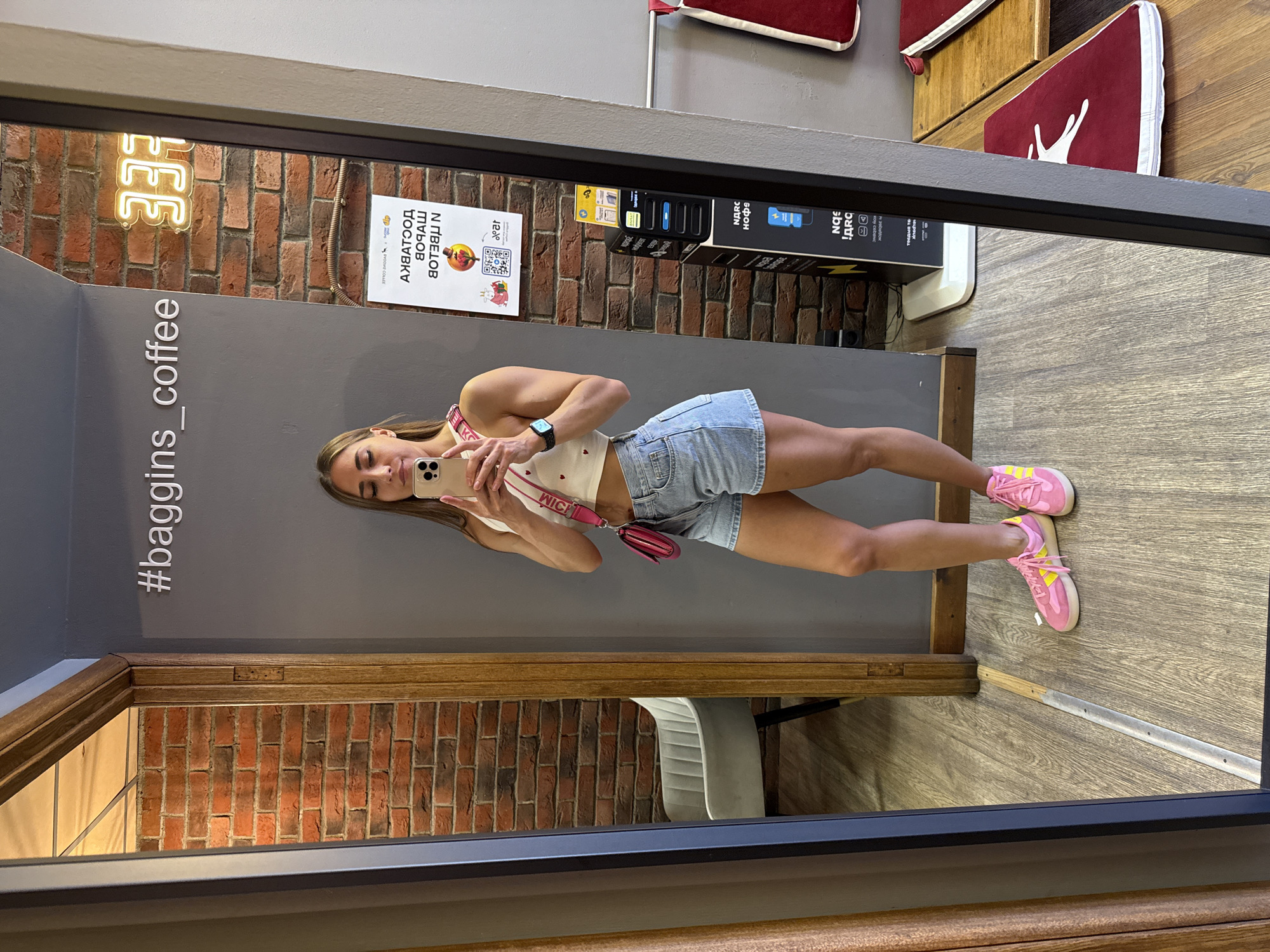 A young woman with blonde hair is taking a mirror selfie in a coffee shop. She is wearing a white crop top with red hearts, denim shorts, and pink sneakers with yellow accents. She has a pink cross-body bag. The coffee shop has exposed brick walls and a gray wall with the hashtag "#baggins_coffee" and a neon sign that says "333". There is a charging station and a table with red cushions visible in the background. The floor is made of light-colored wood. The reflection in the mirror also shows a gray armchair. This image was taken at Baggins Coffee on Bolshaya Morskaya Street in Saint Petersburg, Russia.