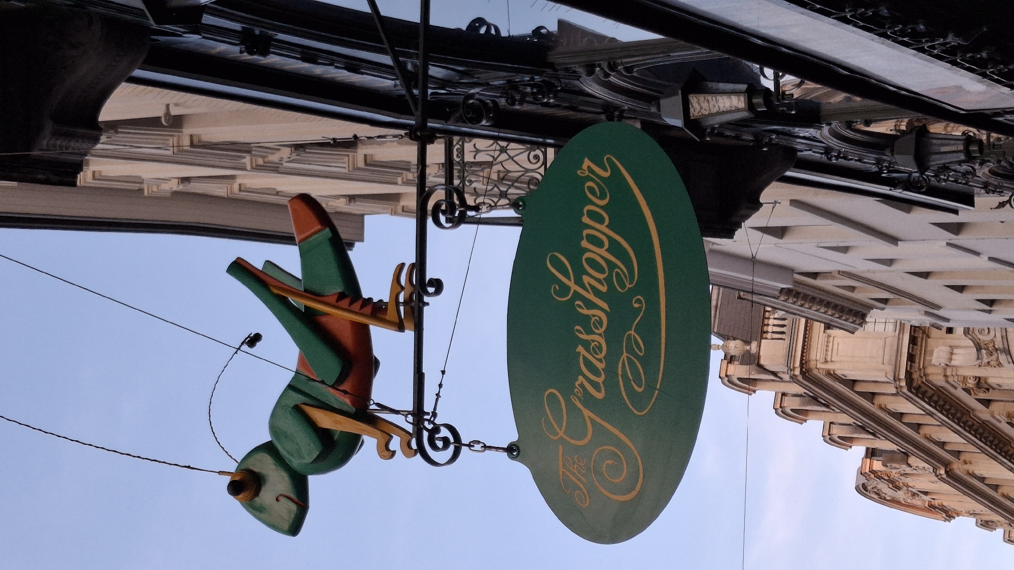 An outdoor, low-angle, full-shot photograph captures a whimsical, green, oval-shaped sign hanging from a building in the city of Unknown, Unknown. The sign, illuminated by the afternoon sun, displays the words "The Grasshopper" in ornate, yellow, cursive lettering. Above the sign, a vibrant green and orange grasshopper sculpture is suspended by wires, its antennae and legs adding to its playful appearance. The grasshopper sculpture is attached to an ornate metal bracket, which in turn is mounted to the facade of an older building. The building is characterized by its intricate architectural details, including decorative stonework, arched windows, and classical moldings. Another building, to the left, exhibits a more modern, cream-colored facade with evenly spaced windows. The sky above is a clear, pale blue.