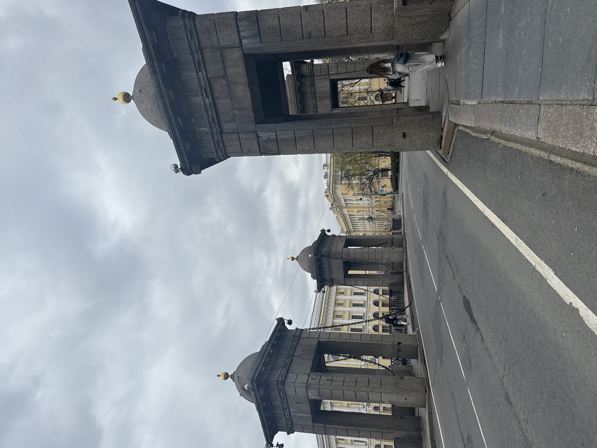The image depicts a street scene in Saint Petersburg, Russia. The architectural elements consist of a series of stone structures or columns with domed tops, possibly part of a bridge or entrance. Chains connect the columns. A road runs between the structures, marked with a white line. Buildings with classical facades are visible in the background. On the right side of the frame, a sidewalk is present, with one or two pedestrians walking along it. The sky is overcast.